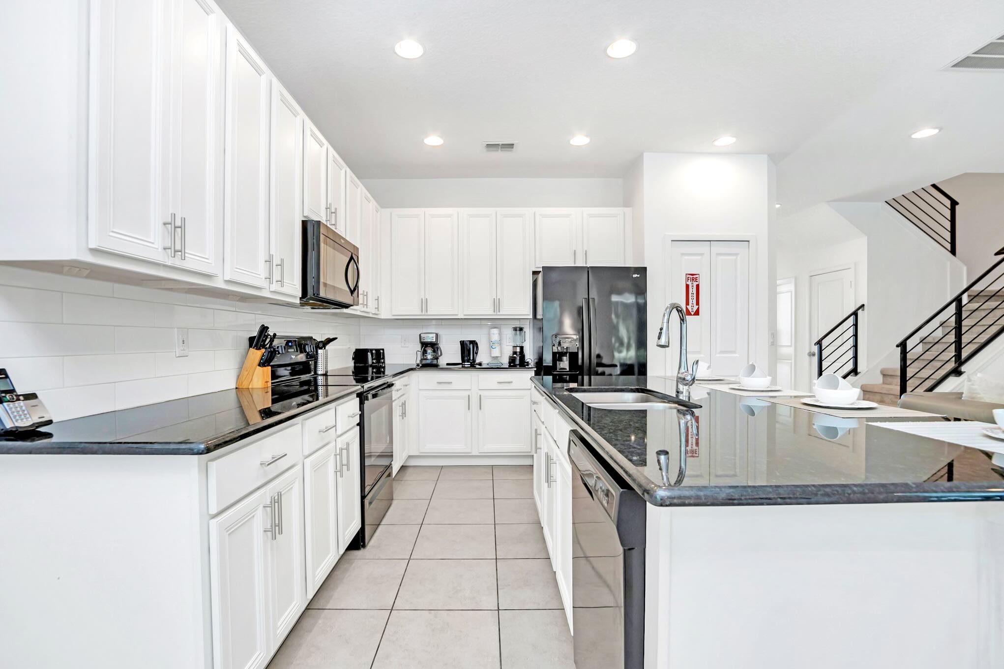 Spacious modern kitchen featuring white shaker cabinets, black granite countertops, and black appliances including a refrigerator, oven, and microwave. A large island with a sink is in the foreground, and tile flooring extends throughout.