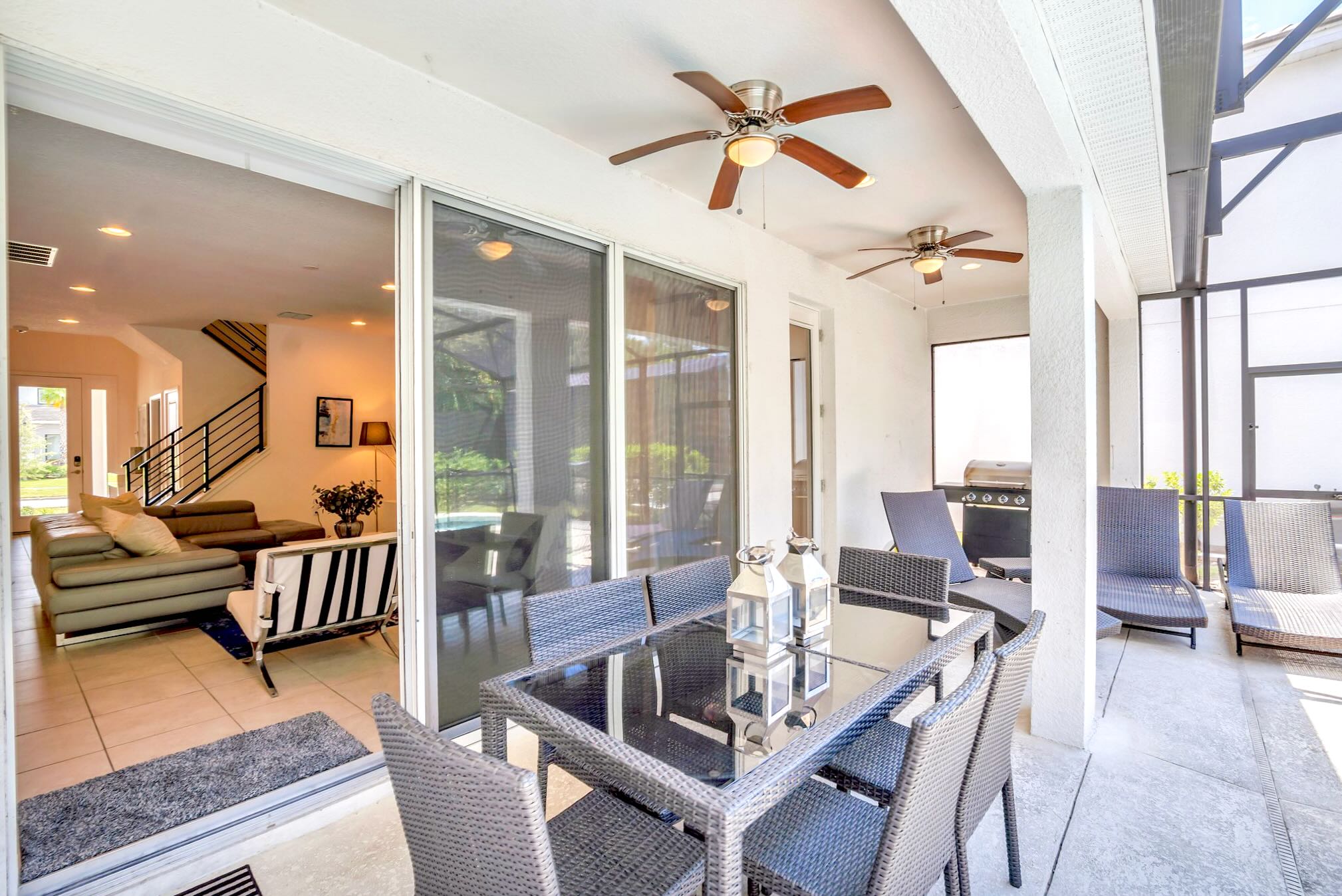 A covered outdoor patio features a glass-top dining table with wicker chairs and two ceiling fans, with a BBQ grill and lounge chairs visible beyond. Sliding glass doors connect to the interior living space.