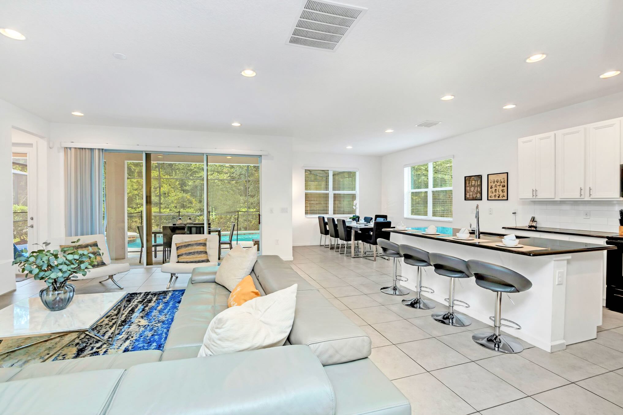 Bright open-concept living space with a large gray sectional sofa, a white coffee table, and a blue and black rug, leading to a dining area and kitchen island with bar stools. Sliding glass doors open to an outdoor pool area.