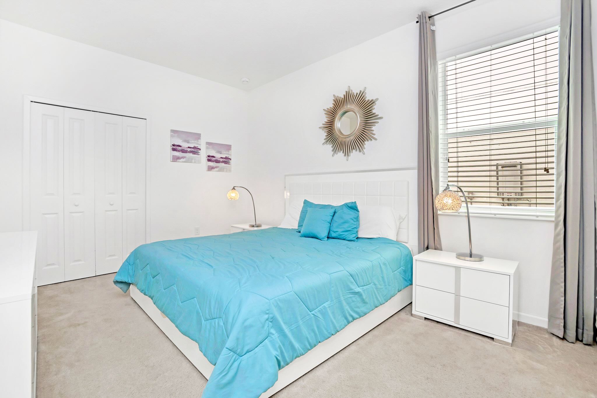 Serene bedroom featuring a white upholstered headboard, a large bed with bright blue bedding, two white nightstands with unique lamps, and a window with blinds and gray curtains.