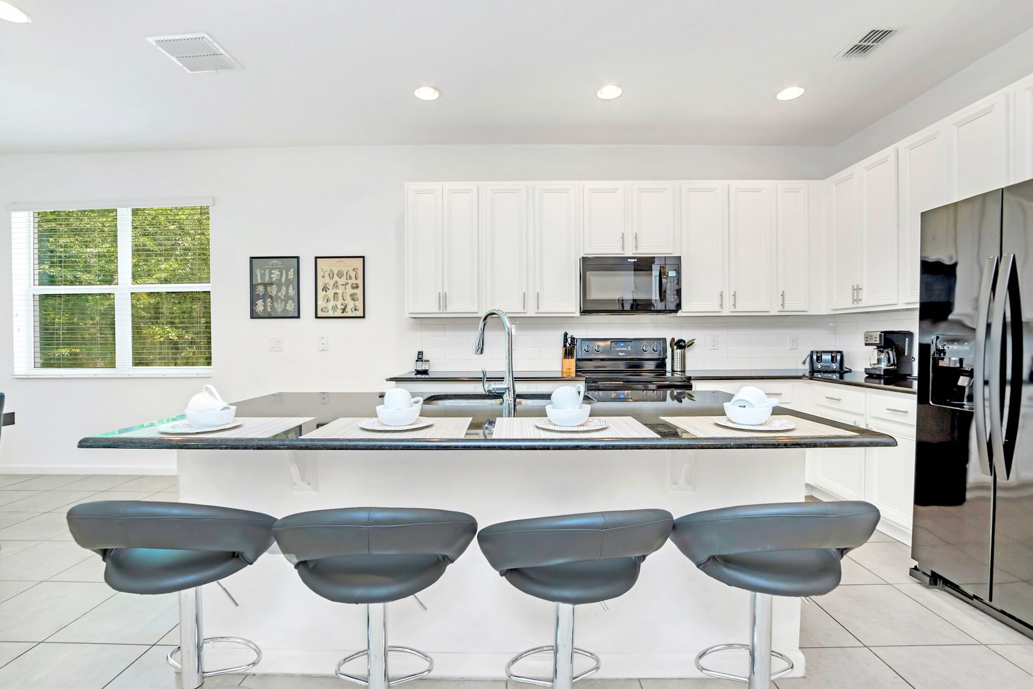 Modern kitchen island with a dark countertop and four black leather bar stools, set with white dishes. White shaker cabinets and black appliances are visible in the background, with a window overlooking greenery.