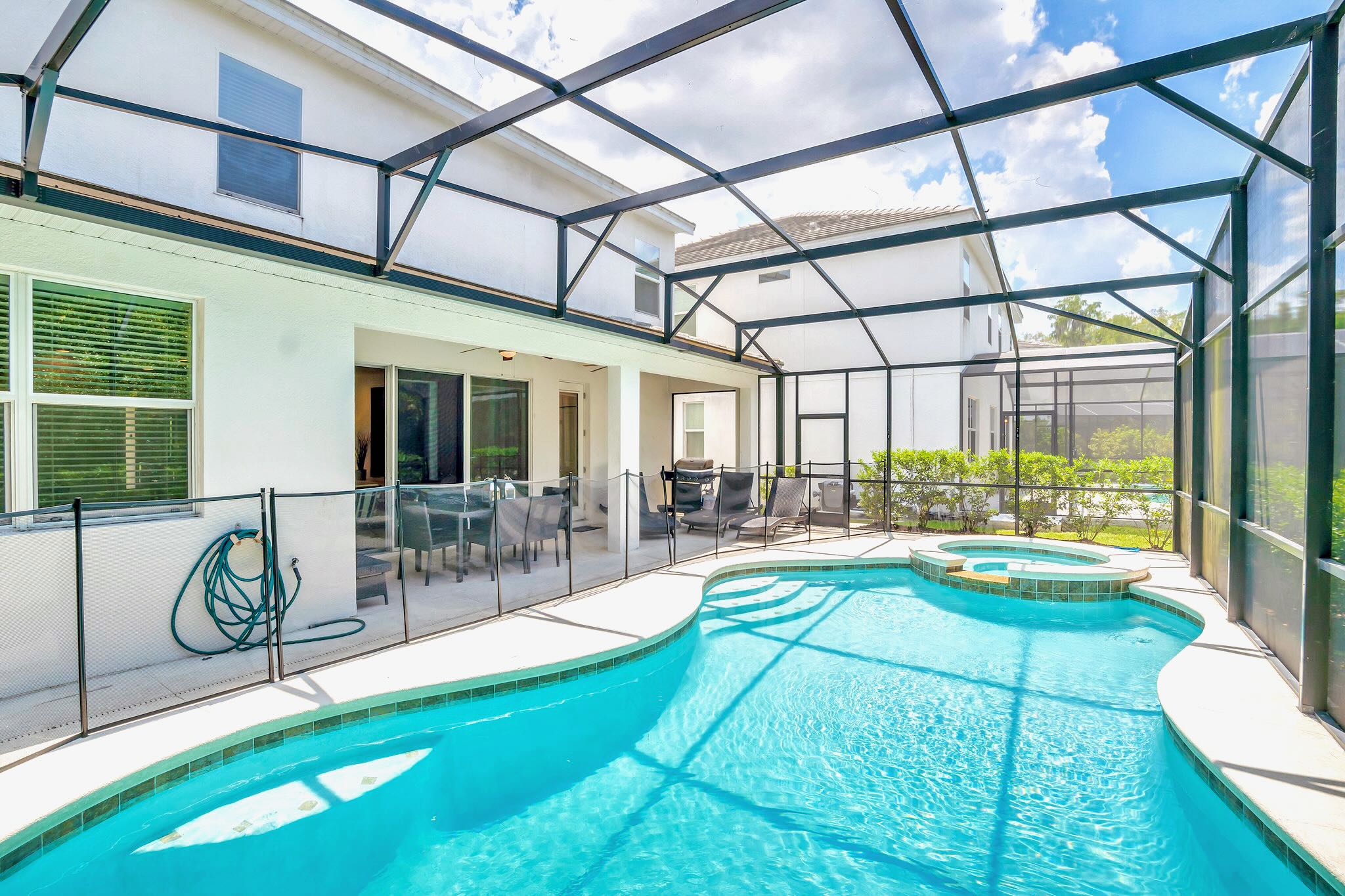 The exterior back of the house features a private swimming pool and hot tub within a screened enclosure, with a covered patio area furnished with outdoor seating and lounge chairs. A child safety fence surrounds the pool.