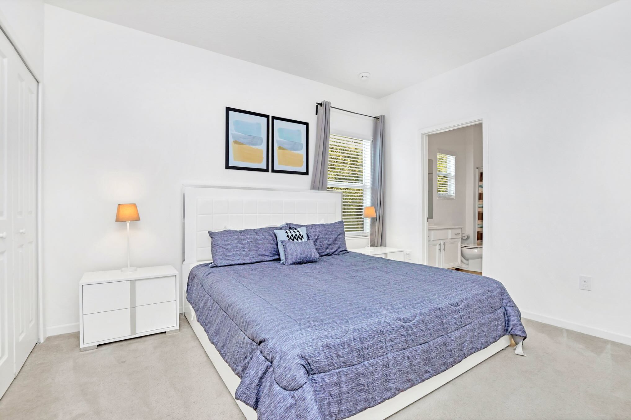 Bright bedroom with a white upholstered headboard, a large bed with blue bedding, two white nightstands with lamps, and a window with blinds and gray curtains.