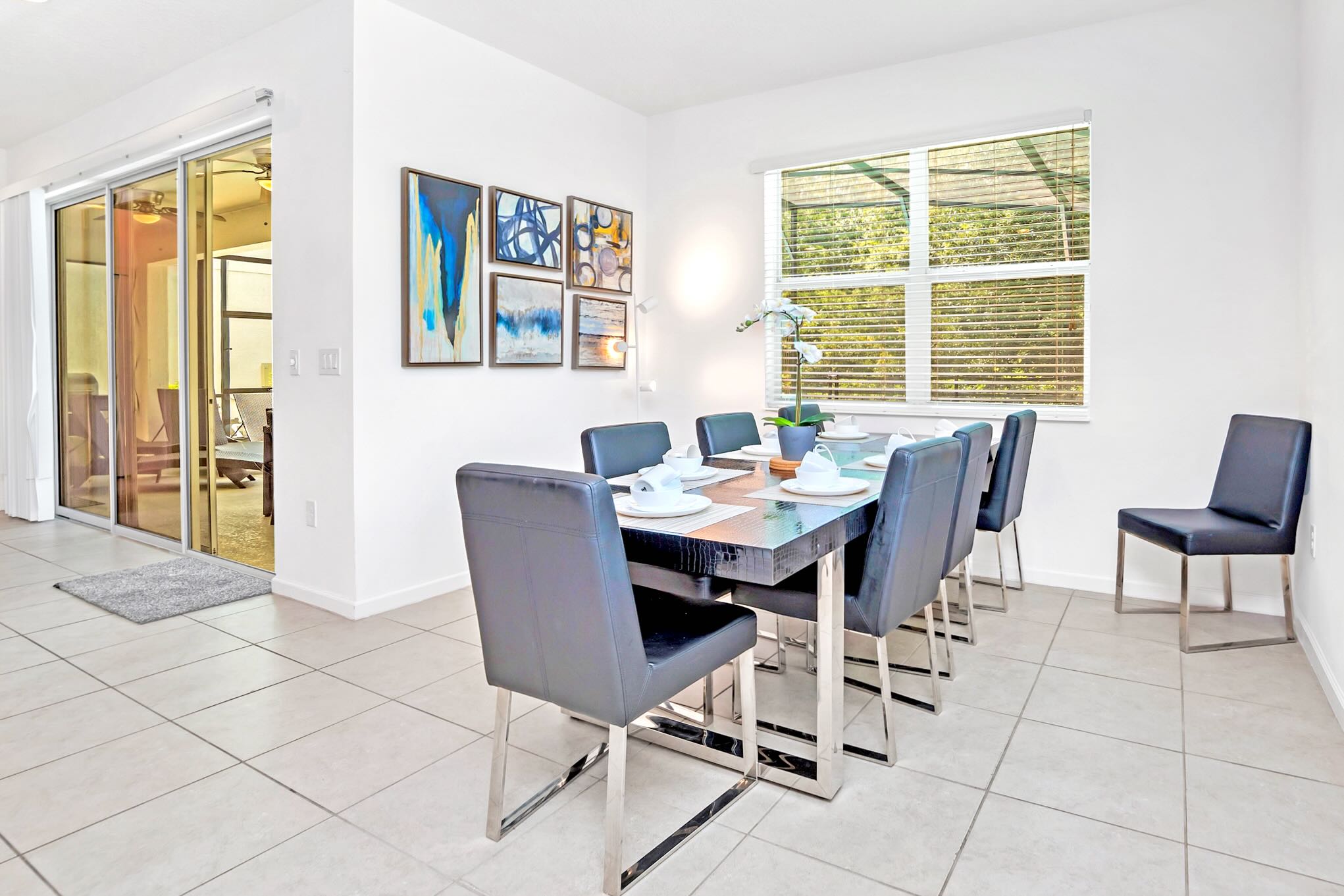 Modern dining room with a dark rectangular table, six black leather chairs with chrome legs, and a set of abstract art pieces on the white wall. A large window with blinds overlooks greenery, and a sliding glass door leads to another room.