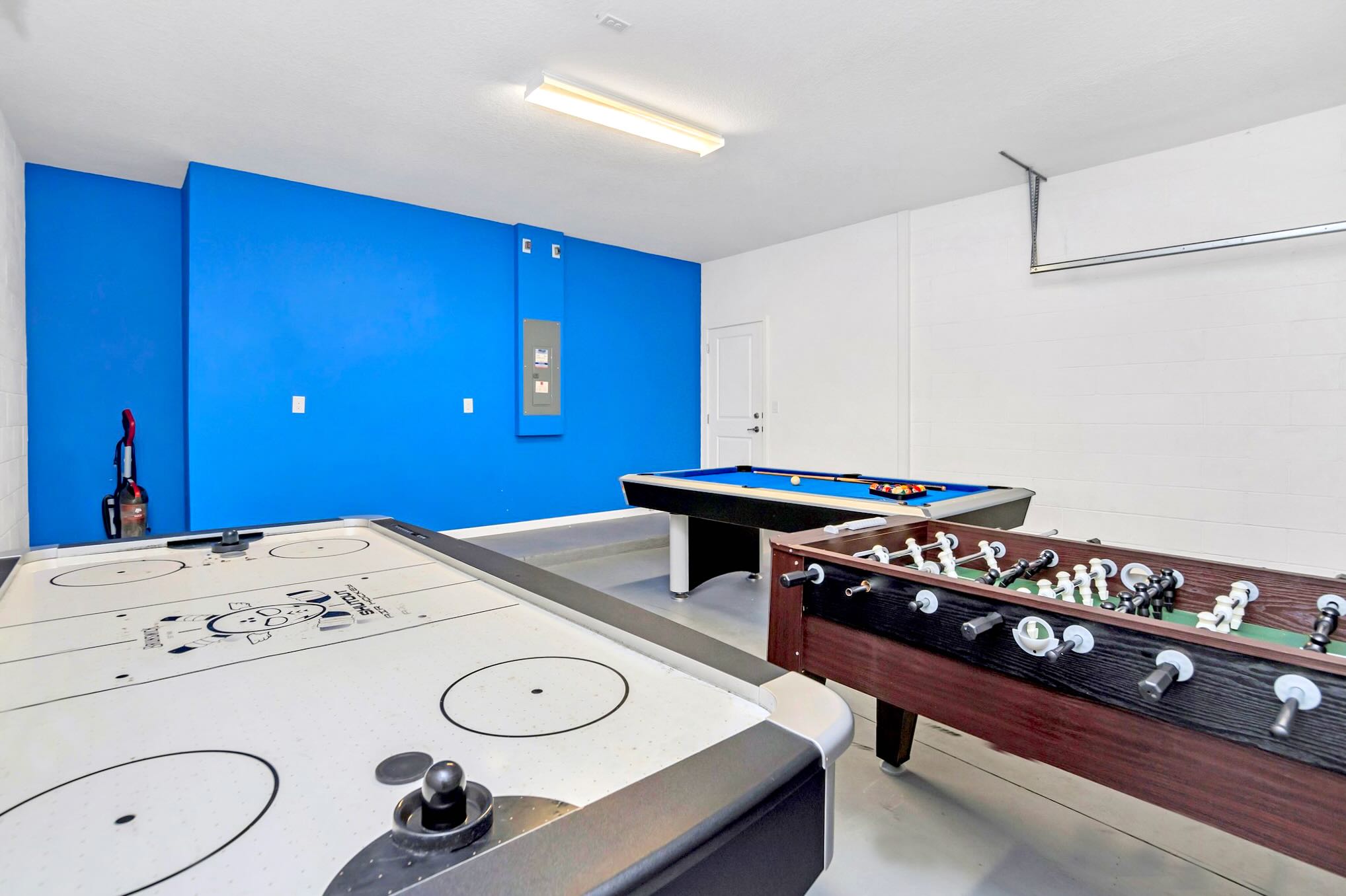 A vibrant game room features an air hockey table, a foosball table, and a pool table, set against a bright blue accent wall and white walls. The floor is a light gray concrete.