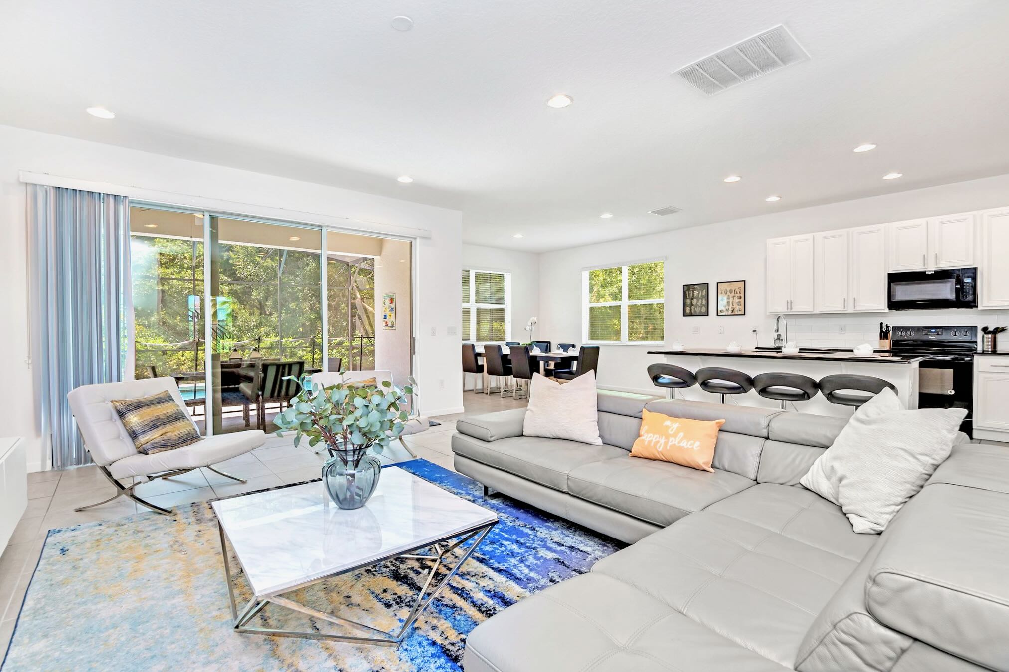 An expansive open-concept living area with light tile flooring connects to a dining area and kitchen, featuring a large gray sectional sofa and sliding glass doors leading to an outdoor patio. Natural light fills the space.