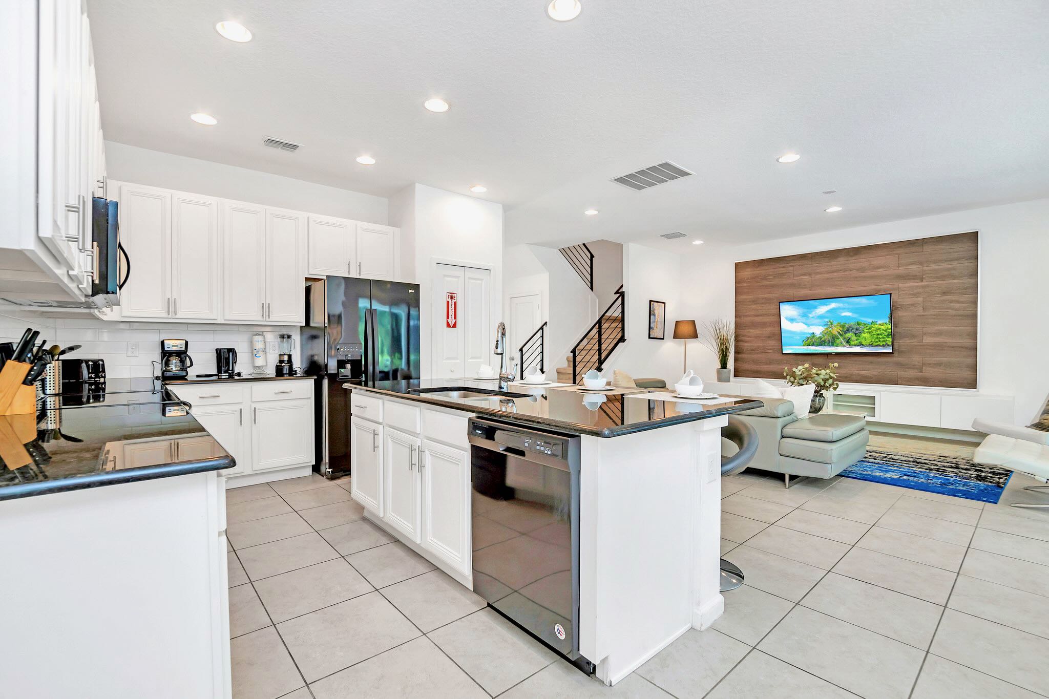 A modern white kitchen with black granite countertops and stainless steel appliances features a large island with bar seating, seamlessly opening into the living room area. Recessed lighting illuminates the space.