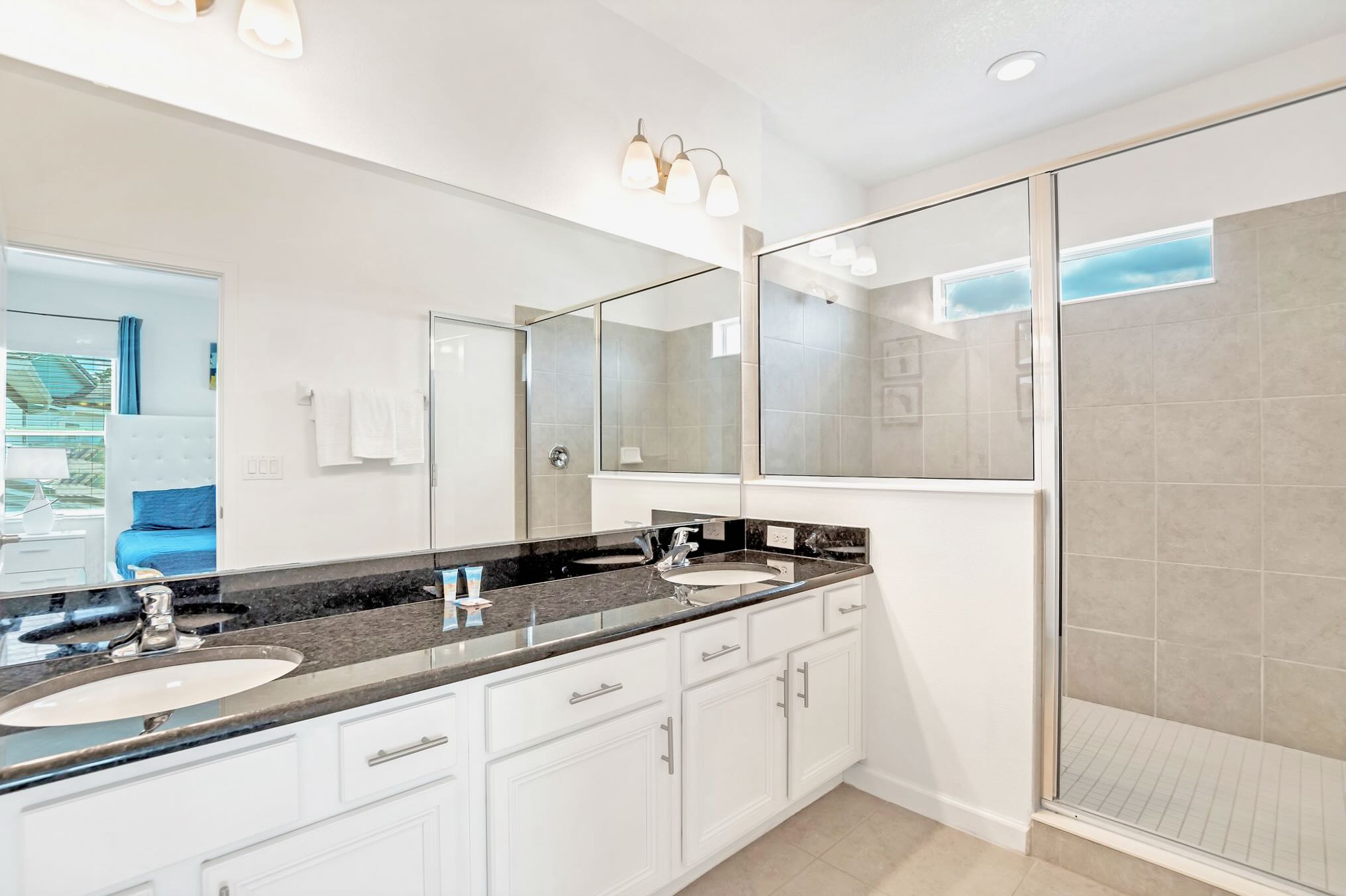 Modern bathroom featuring a black granite double vanity with two sinks, white cabinets, a large mirror, and a spacious walk-in shower with light gray tiled walls.