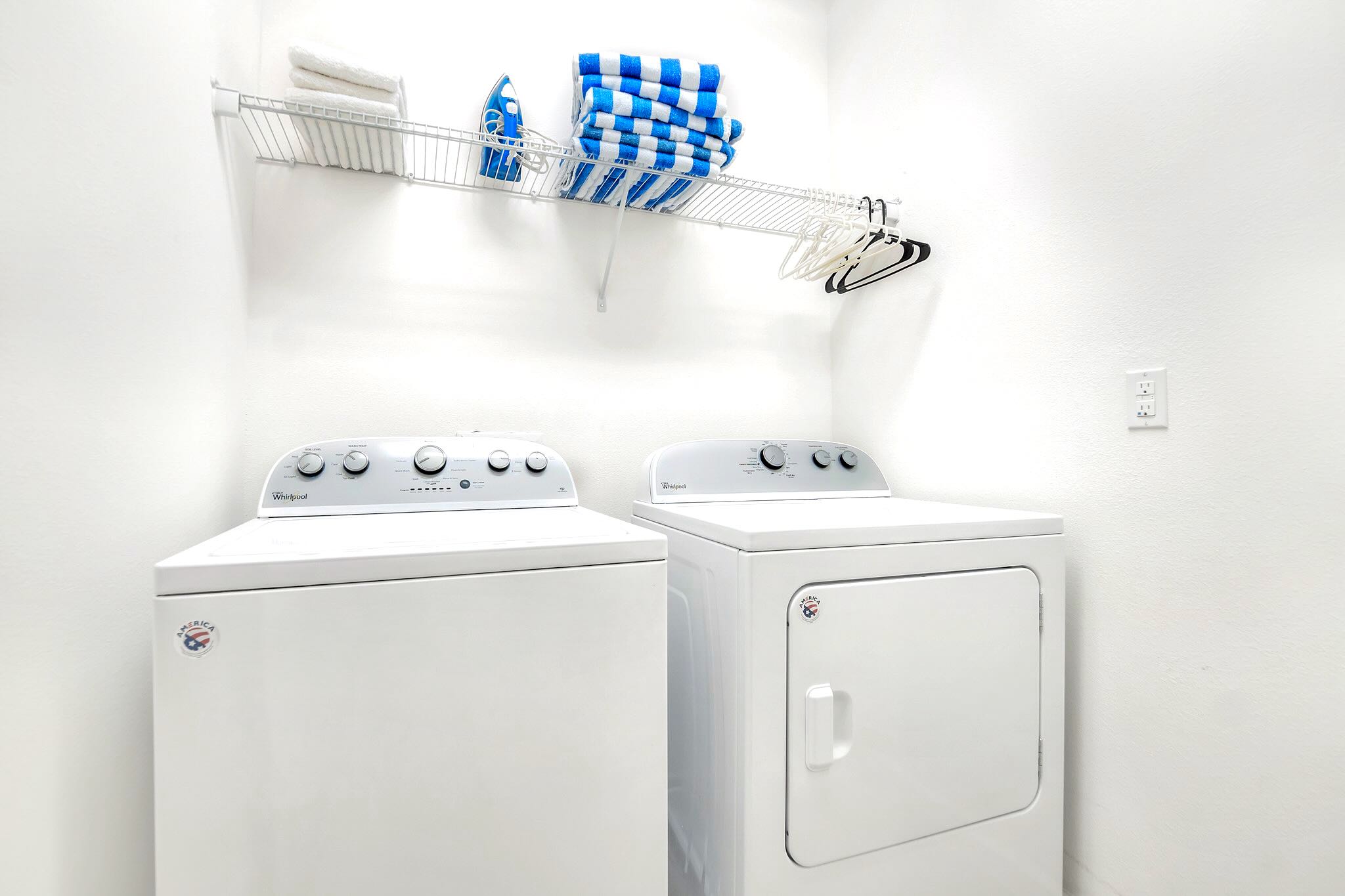 Functional laundry room equipped with a white top-loading washing machine and a front-loading dryer, a wire shelf with towels and an iron, and white walls.