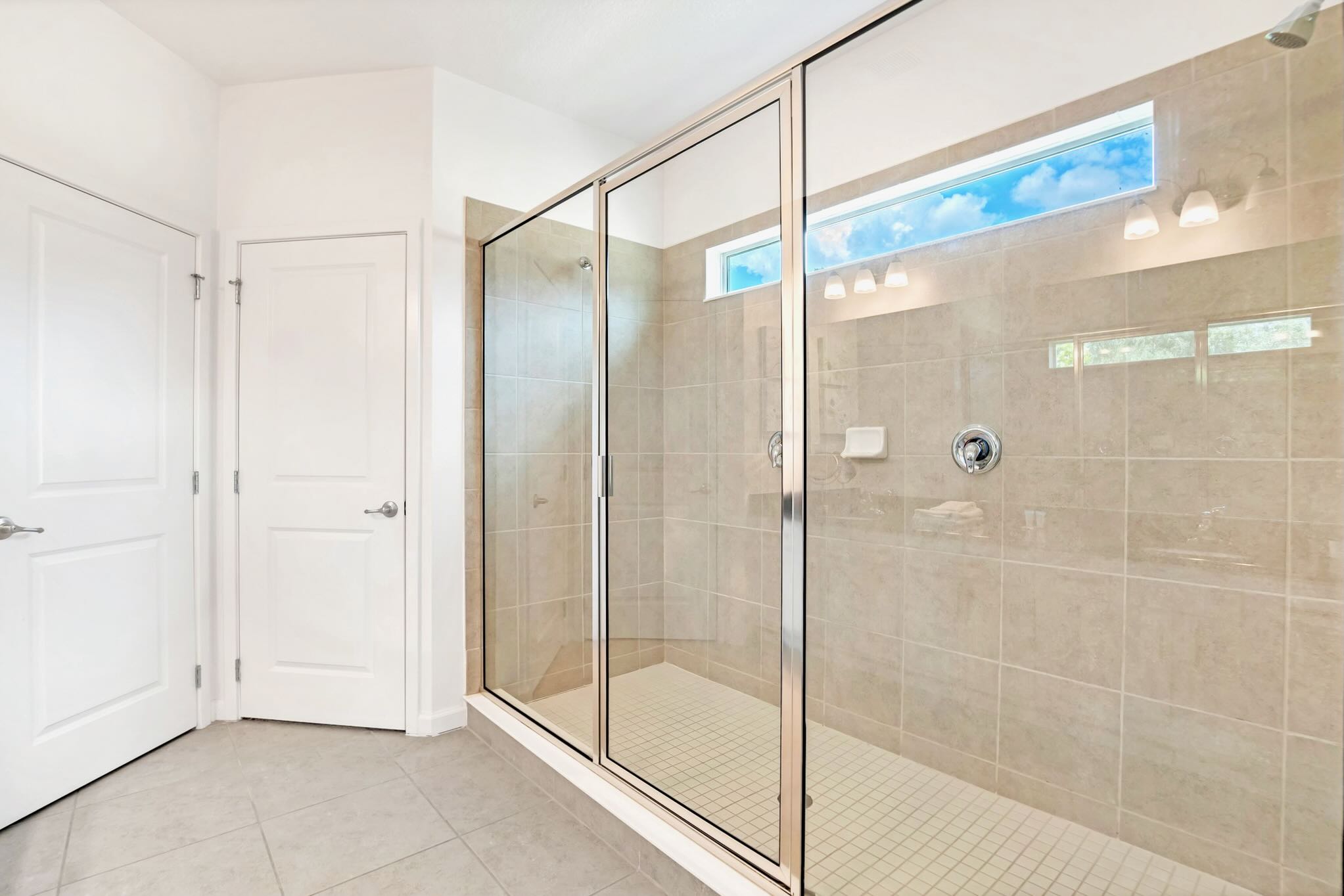 Spacious walk-in shower with light beige tiled walls and floor, enclosed by clear glass panels. A high horizontal window allows natural light, and white doors lead to other areas of the bathroom.