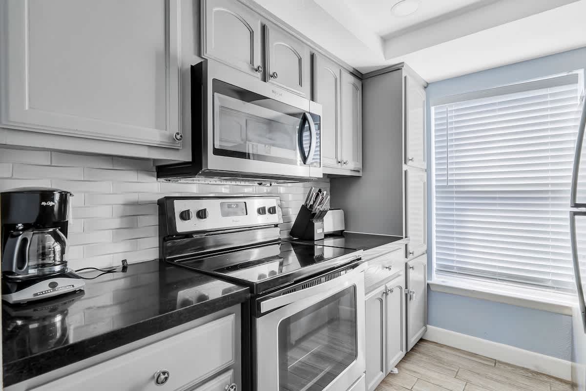 View of the kitchen's cooking area, featuring a stainless steel electric range with an over-the-range microwave. Black countertops, gray shaker cabinets, and white textured subway tile backsplash complete the modern look.