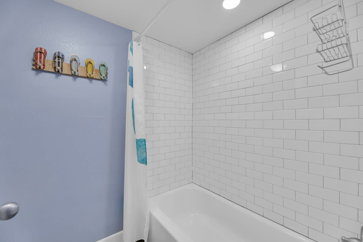 A clean bathroom featuring a white subway-tiled shower/tub combo with a white shower curtain and a light blue painted wall adorned with colorful flip-flop decor. A metal shower caddy is visible in the corner.