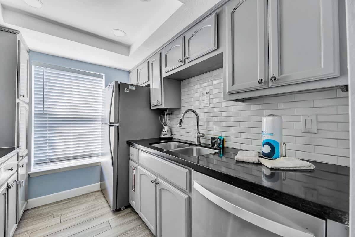 Close-up view of the kitchen sink area with a double basin stainless steel sink, black countertop, and white textured subway tile backsplash. Gray shaker cabinets are above and below, with a stainless steel dishwasher visible.