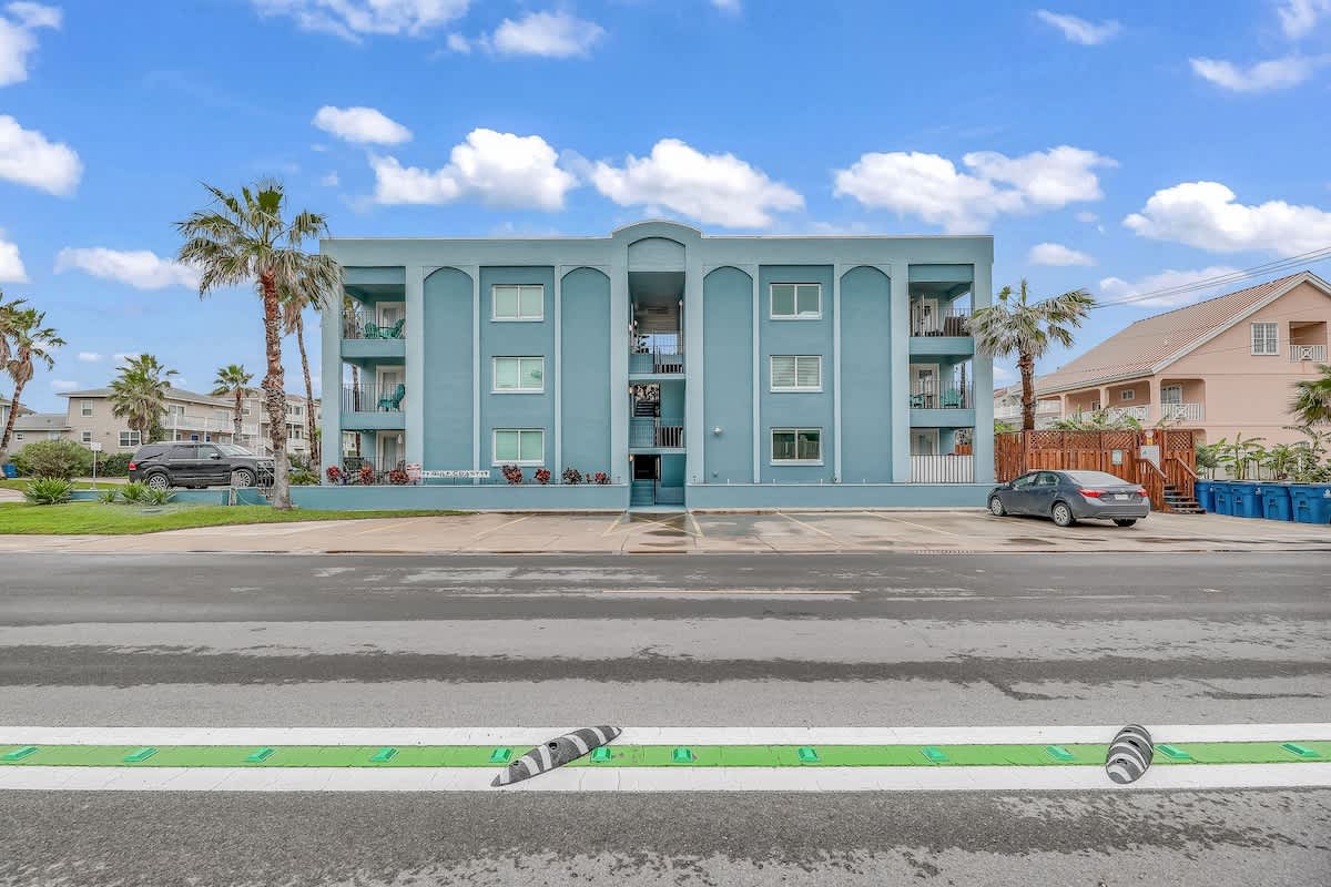 The front exterior of a three-story light blue apartment building with balconies and a central outdoor staircase, seen from across a street with a parking area. Palm trees frame the building, and a car is parked on the right.