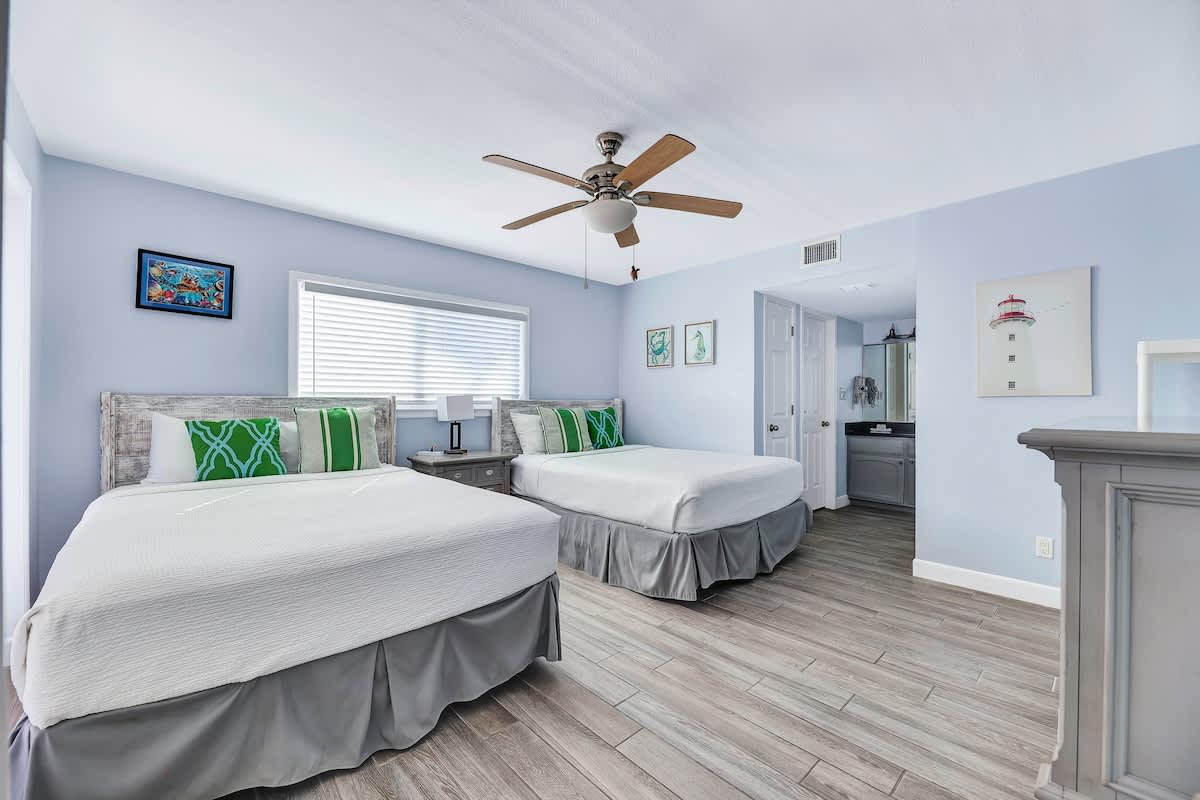 Bright bedroom with light blue walls and wood-look tile flooring, featuring two queen beds with gray bed skirts and green and white striped pillows. A ceiling fan and a window with blinds are visible, along with coastal artwork.