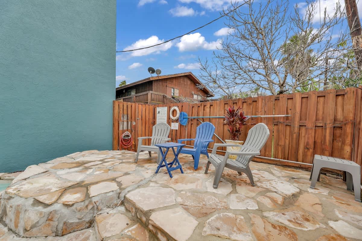 An outdoor seating area with flagstone paving, featuring three Adirondack-style chairs and a small table in shades of blue and gray. A wooden fence encloses the space, with some greenery and a house visible in the background.