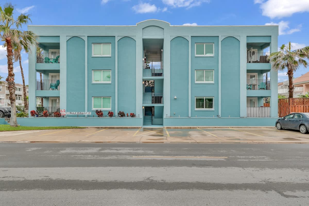 The front view of a three-story light blue apartment building with multiple balconies, a central outdoor staircase, and a parking lot in front. Palm trees flank the building, and a car is parked on the right.