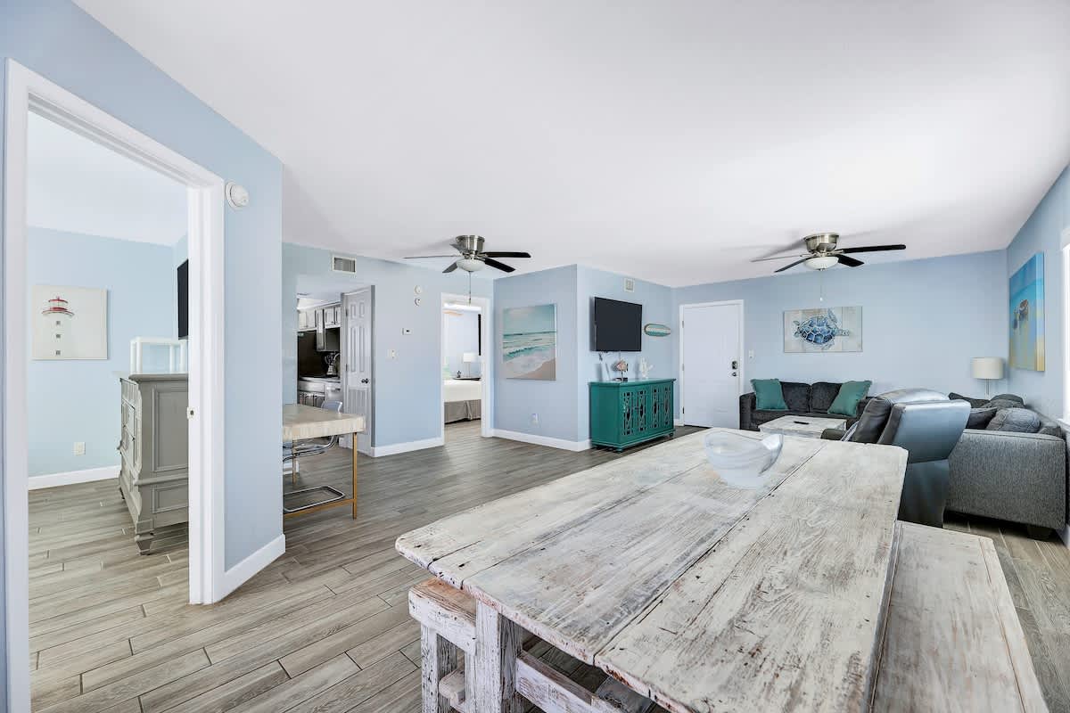 Wide view of the open living and dining space featuring light blue walls, wood-look tile flooring, a large distressed white dining table, and a gray sofa. A doorway leads to a kitchen area, and another to a bedroom.