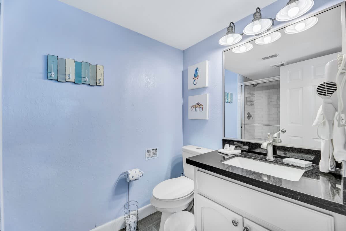 Bathroom with light blue walls, a white toilet, and a vanity with a black countertop and white cabinet. Coastal-themed wall decor and a toilet paper holder are visible, with a shower enclosure to the right.