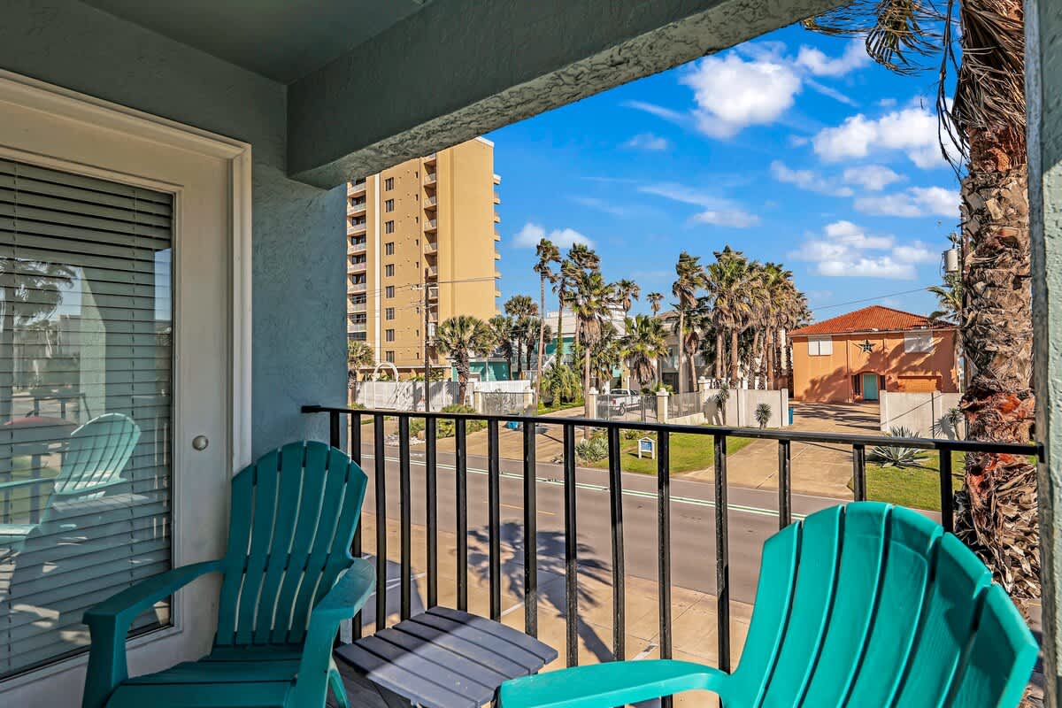 Balcony view from behind two teal Adirondack chairs, offering a clear perspective of the street below, distant buildings, and numerous palm trees under a bright blue sky.