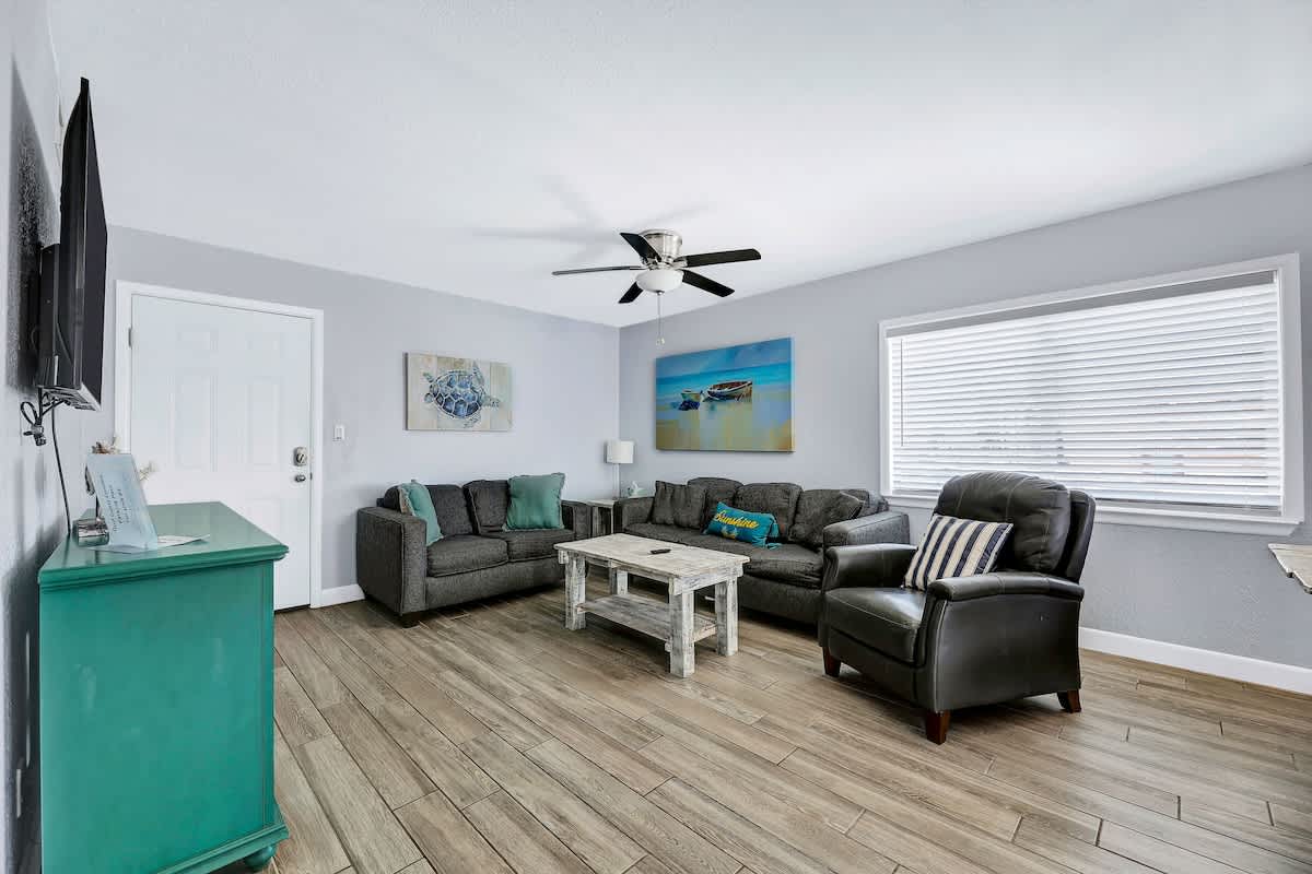 Spacious living room with light gray walls, two gray sofas, a dark leather armchair, and a distressed wooden coffee table, with a large window providing natural light.