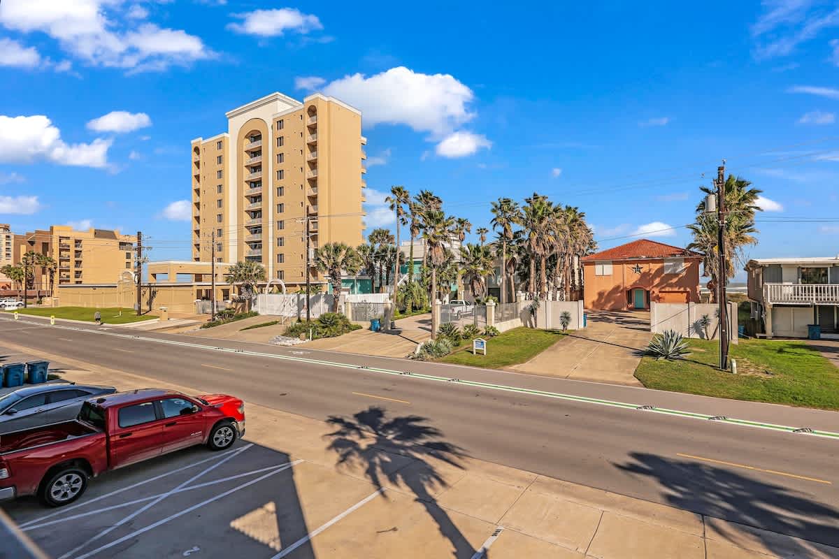 An elevated view of a coastal street with a red pickup truck parked in the foreground, overlooking other buildings and numerous palm trees. A tall, light-colored high-rise building stands prominently in the distance under a blue sky with white clouds.