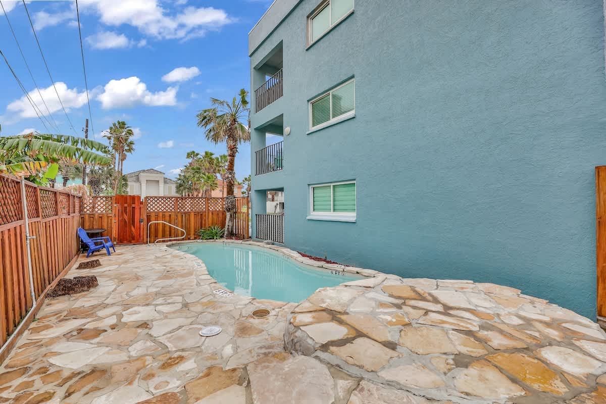 A small, irregularly shaped outdoor swimming pool surrounded by flagstone paving and a wooden fence. The light blue exterior of the building is visible on the left, with palm trees in the background.