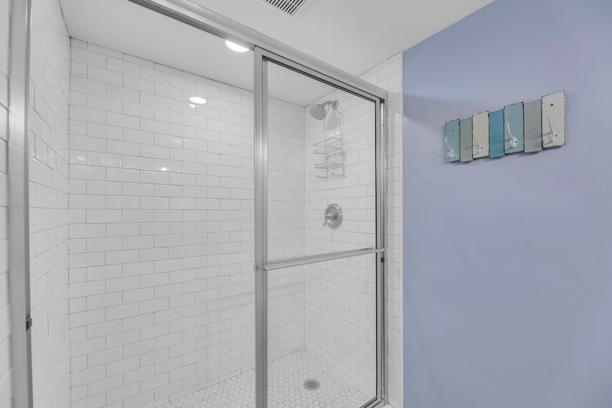 Modern shower enclosure with white subway tiles on the walls and white hexagonal mosaic tiles on the floor. A sliding glass door encloses the shower, and a shower caddy is mounted inside.