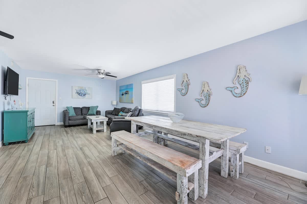 Open concept living and dining area with light blue walls, wood-look tile flooring, a distressed white wooden dining table with benches, and a gray sofa in the living space. Coastal decor includes mermaid wall art and a turquoise console.