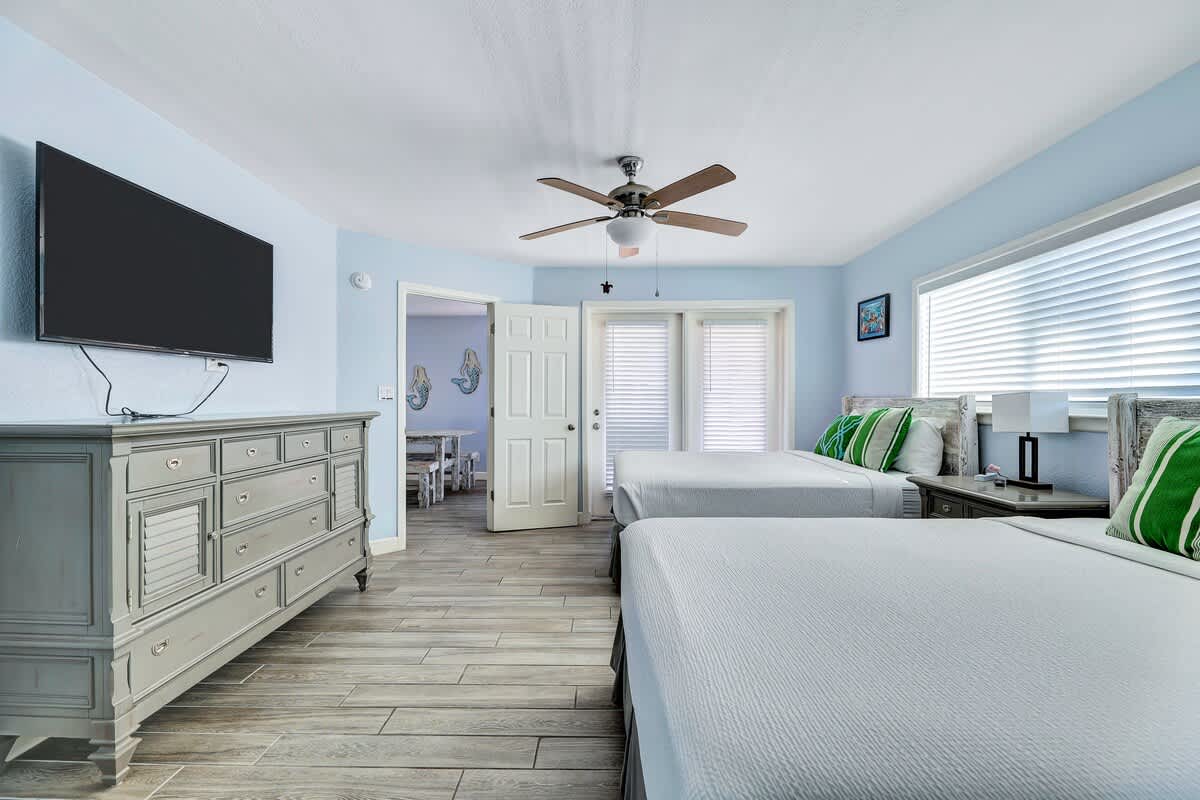 Another angle of the bedroom showing two queen beds, a large gray dresser with a flat-screen TV, and a doorway leading to the living area. Light blue walls and wood-look tile flooring maintain the cohesive coastal theme.