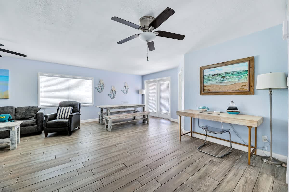 Open-concept living space with light blue walls, featuring a gray sofa and armchair, a rustic dining table with benches, and a modern desk area, all on wood-look flooring.