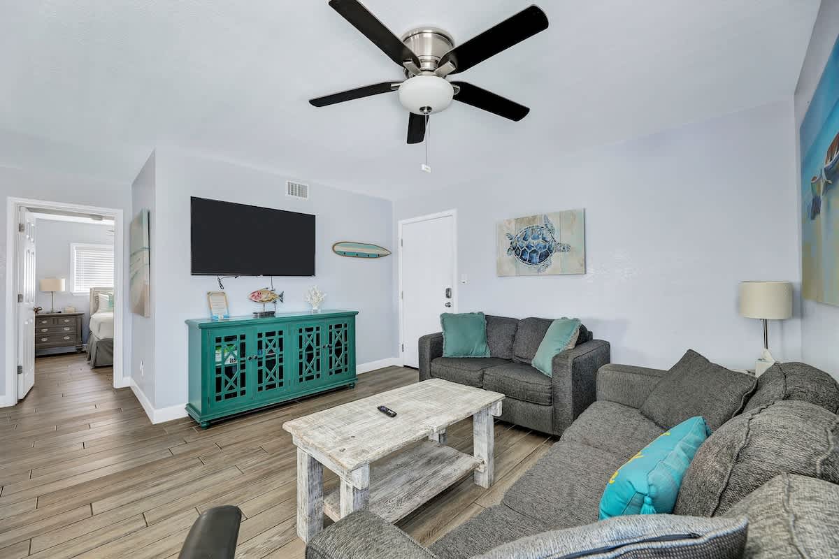 Cozy living room featuring a wall-mounted flat-screen TV above a teal console table, a gray sectional sofa with throw pillows, and a rustic wooden coffee table, all on wood-look flooring.