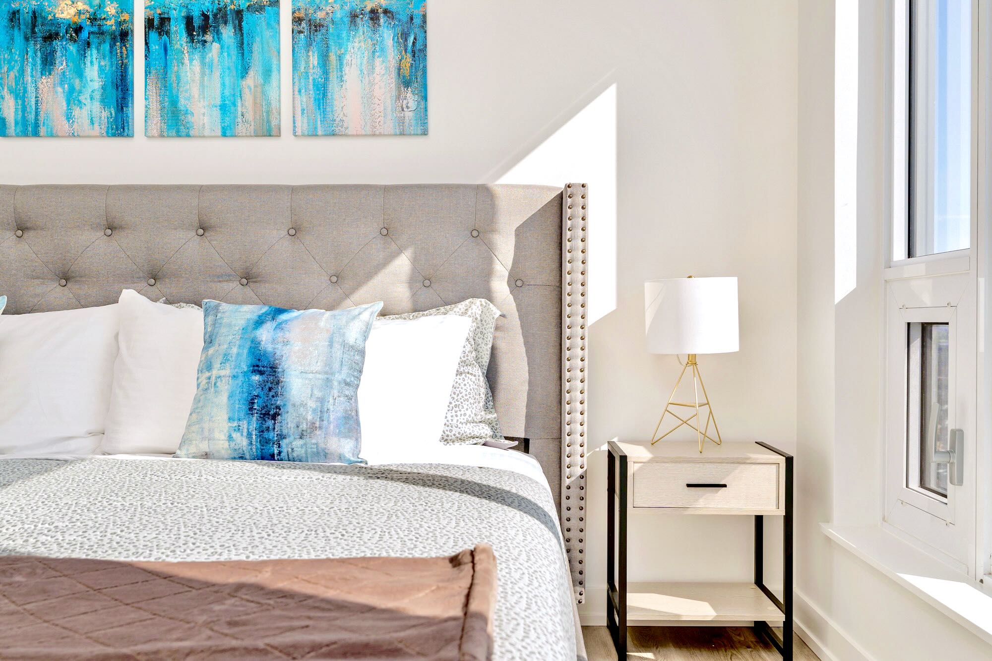A close-up of the bedroom showing the grey tufted headboard, white and blue pillows, a white bedside table with a gold-accented lamp, and a large window letting in natural light.