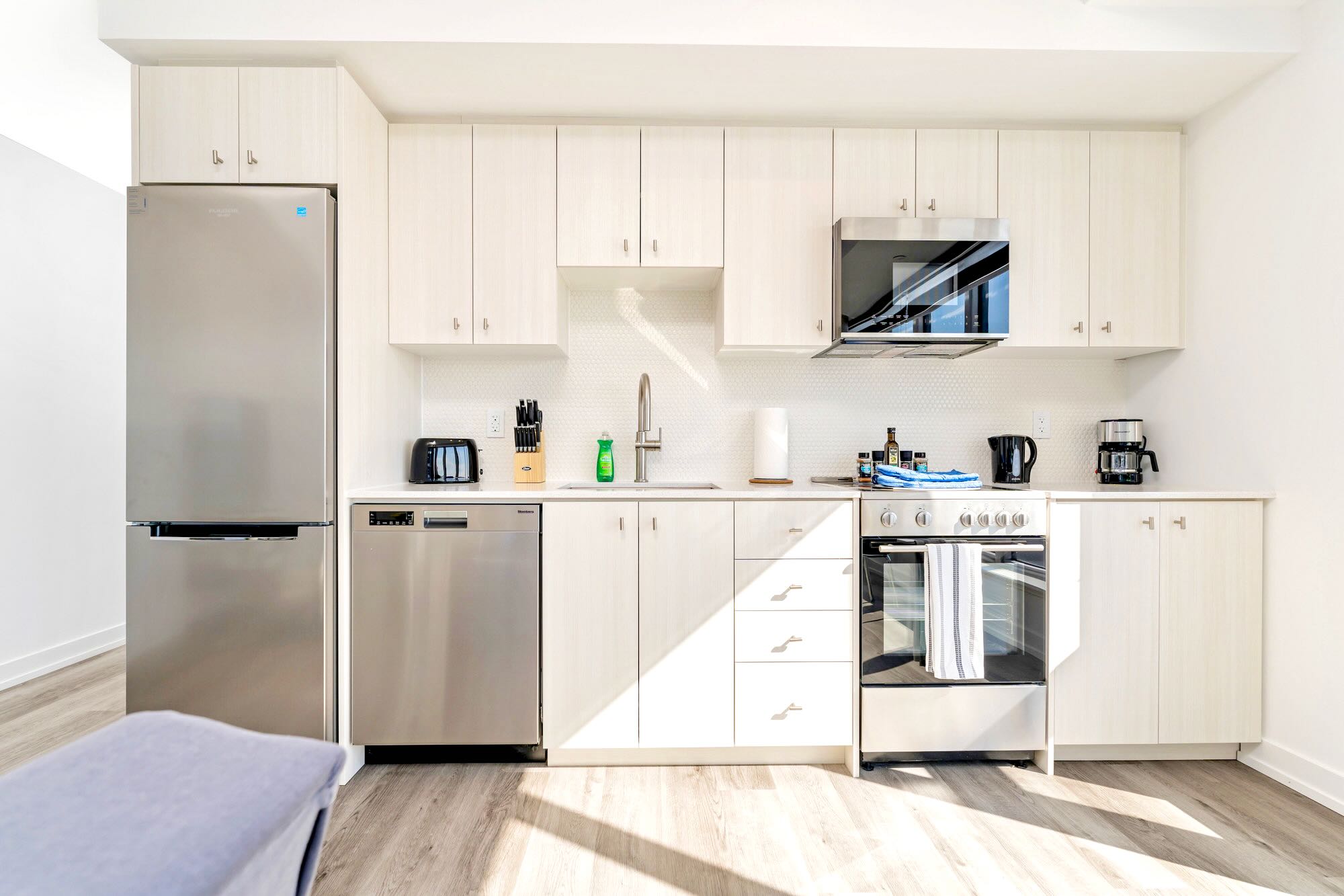 A clean and modern kitchen with light wood-look cabinetry, white subway tile backsplash, and stainless steel appliances including a refrigerator, dishwasher, oven, and microwave, equipped with various kitchen essentials.