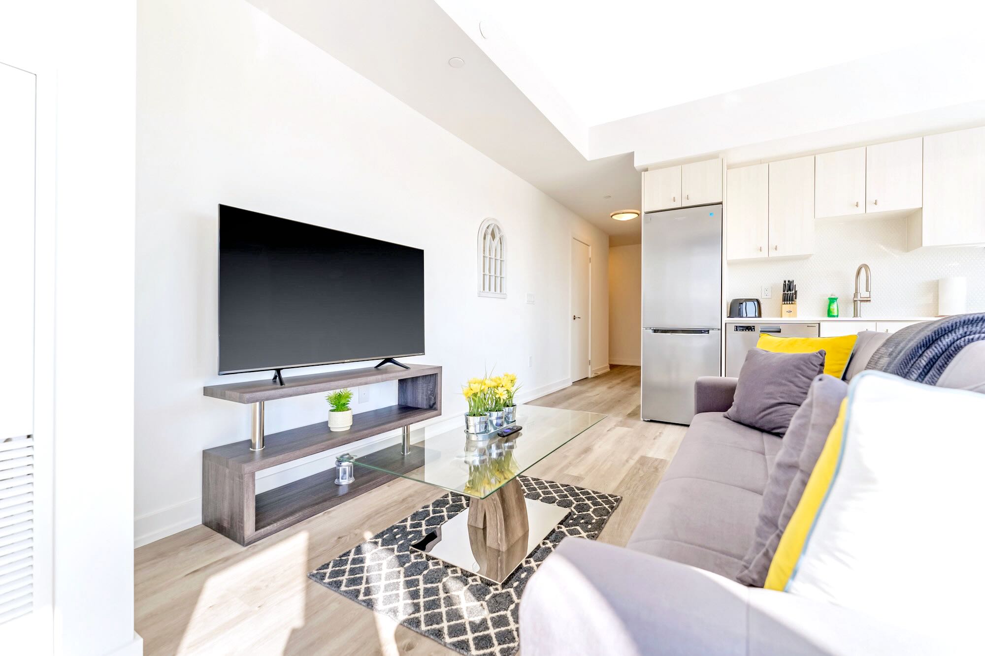 A cozy living room with light wood-look flooring, a grey sofa with yellow and grey throw pillows, a glass coffee table, and a large flat-screen TV on a wooden stand, with a partial view of the kitchen in the background.