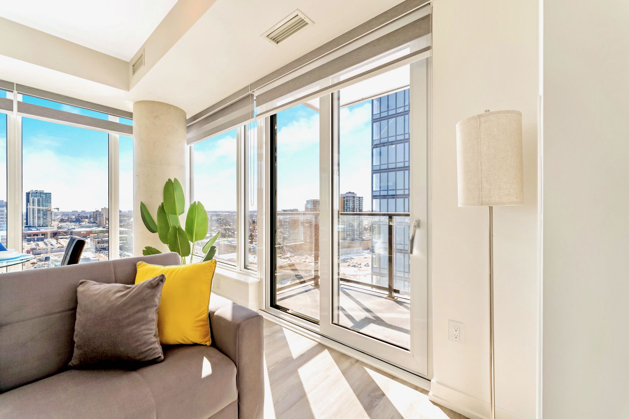 A bright living room corner with a grey sofa and colorful pillows, featuring a tall floor lamp and large sliding glass doors leading to a balcony, offering a glimpse of the city outside.
