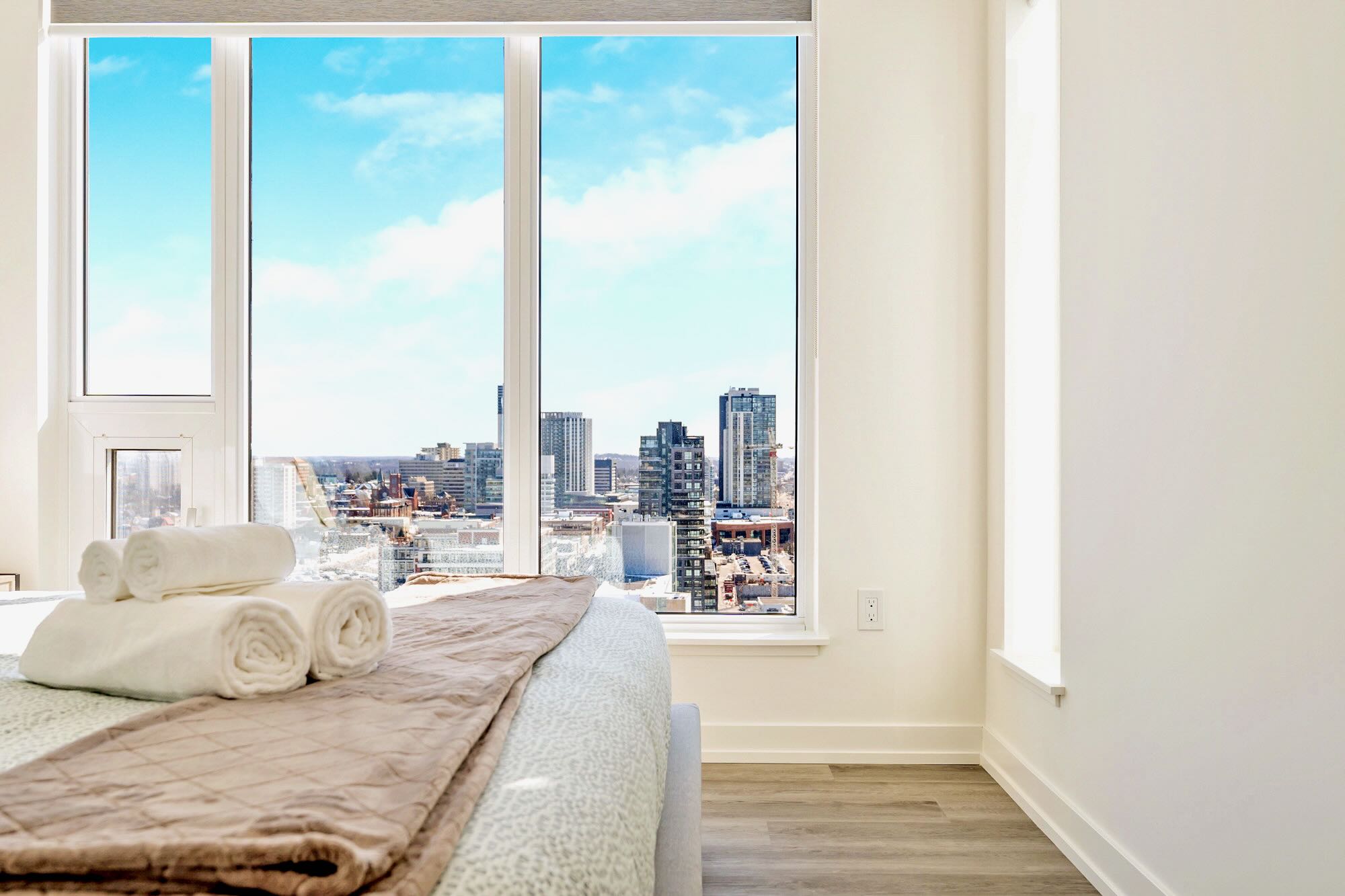A bedroom with a large window offering panoramic city views, showing tall buildings against a blue sky. Rolled white towels are neatly placed on the bed, which has a brown throw blanket.