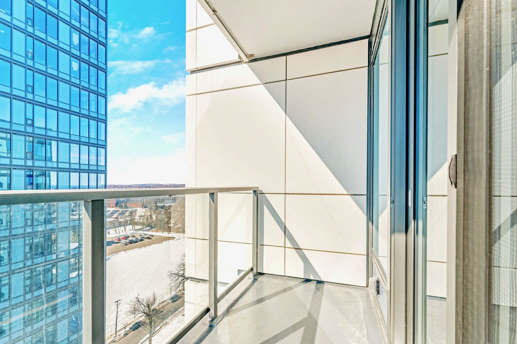 A narrow balcony with a glass railing provides a view of a snow-covered ground below and a modern glass-facade building adjacent, under a bright blue sky.