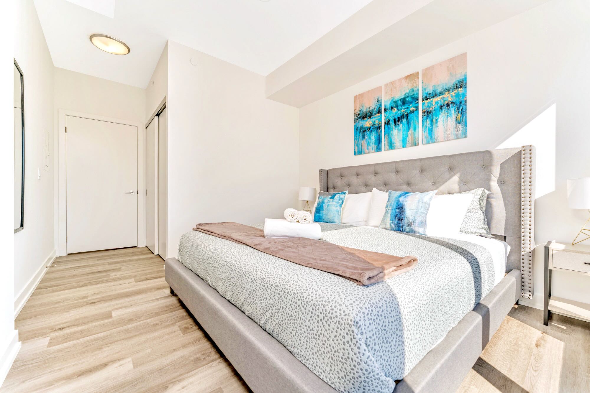 A bright bedroom featuring a large grey upholstered bed with a tufted headboard, light wood-look flooring, and three abstract blue and gold art pieces above the bed. A white door leads to a closet or another room.