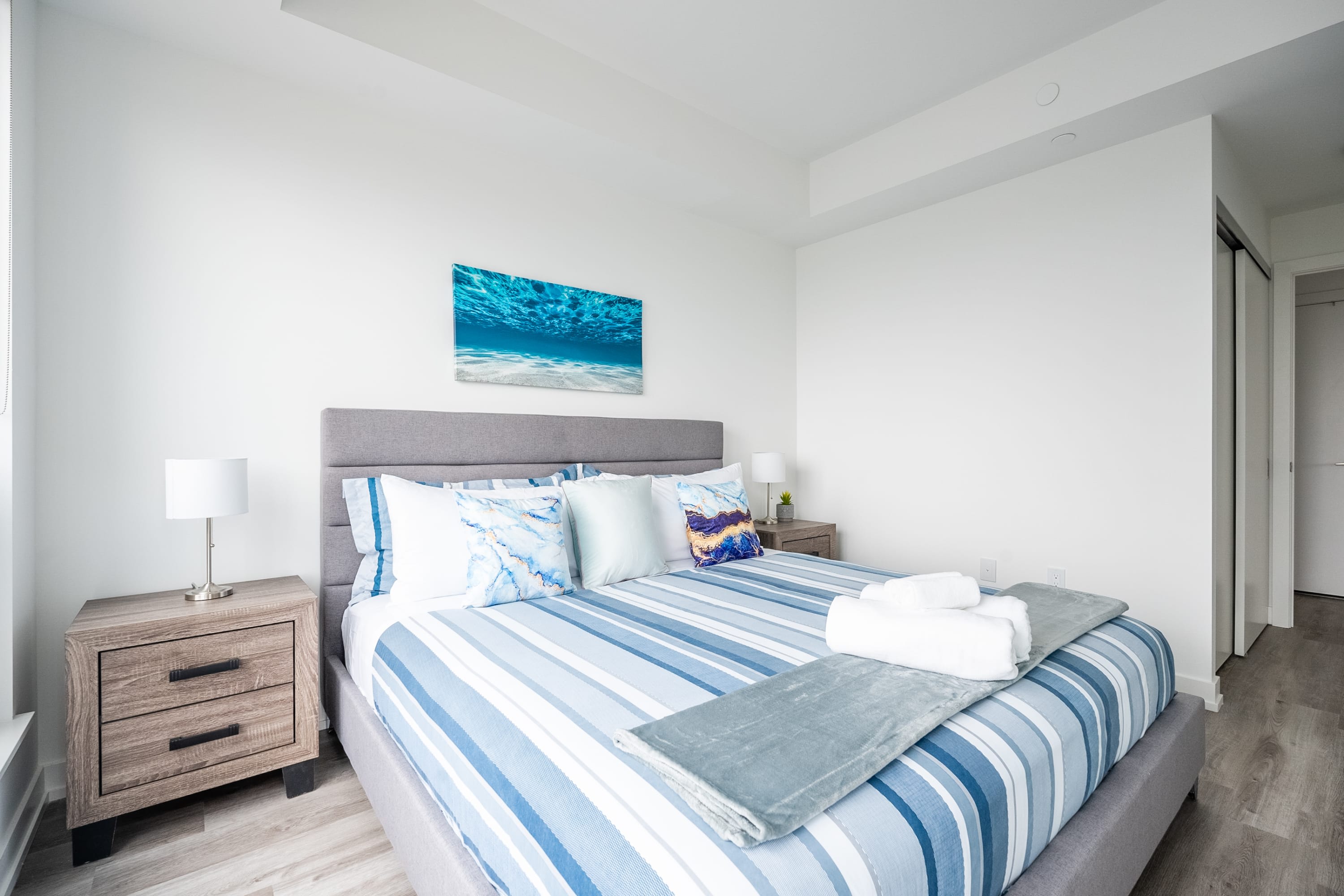 Cozy bedroom featuring a queen-sized bed with a grey upholstered headboard, blue and white striped bedding, and two wooden nightstands with white lamps, complemented by an underwater-themed artwork above the bed.