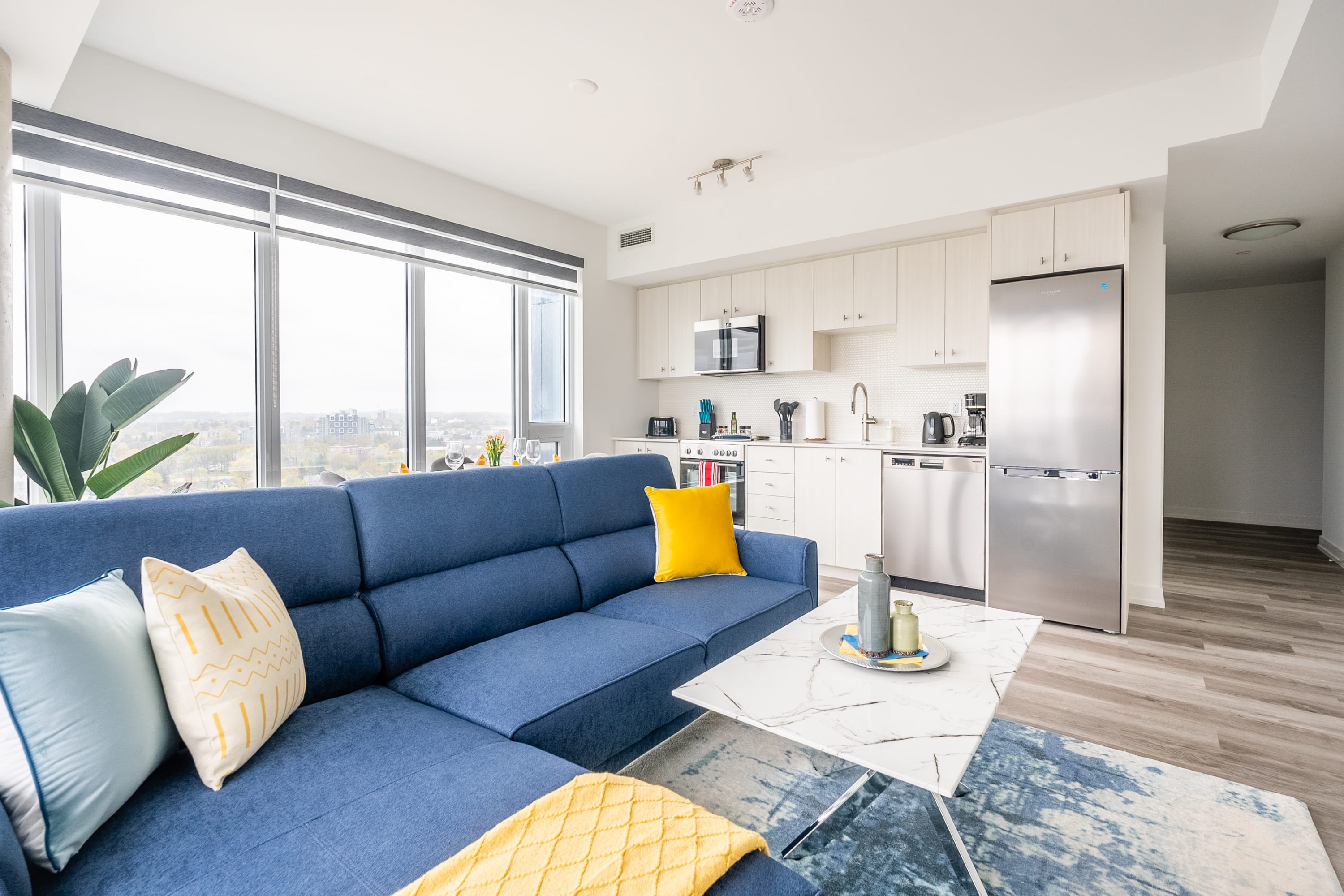 Open-concept living space featuring a blue sectional sofa with yellow and white pillows, a white marble coffee table, and a modern kitchen with light wood cabinets and stainless steel appliances.