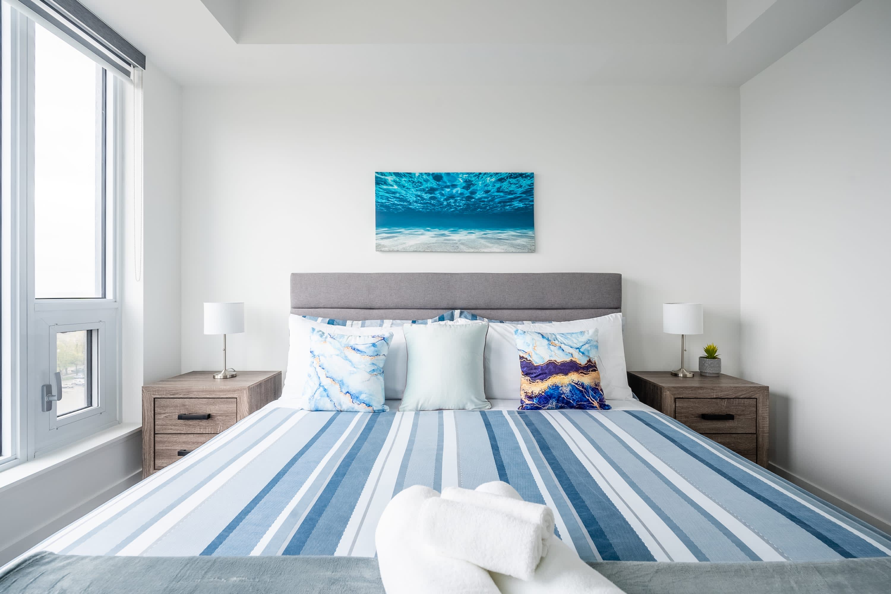 Bright bedroom with a queen bed dressed in blue and white striped linens, flanked by two wooden nightstands with white lamps, and a large window providing natural light, with an ocean-themed artwork centered above the bed.