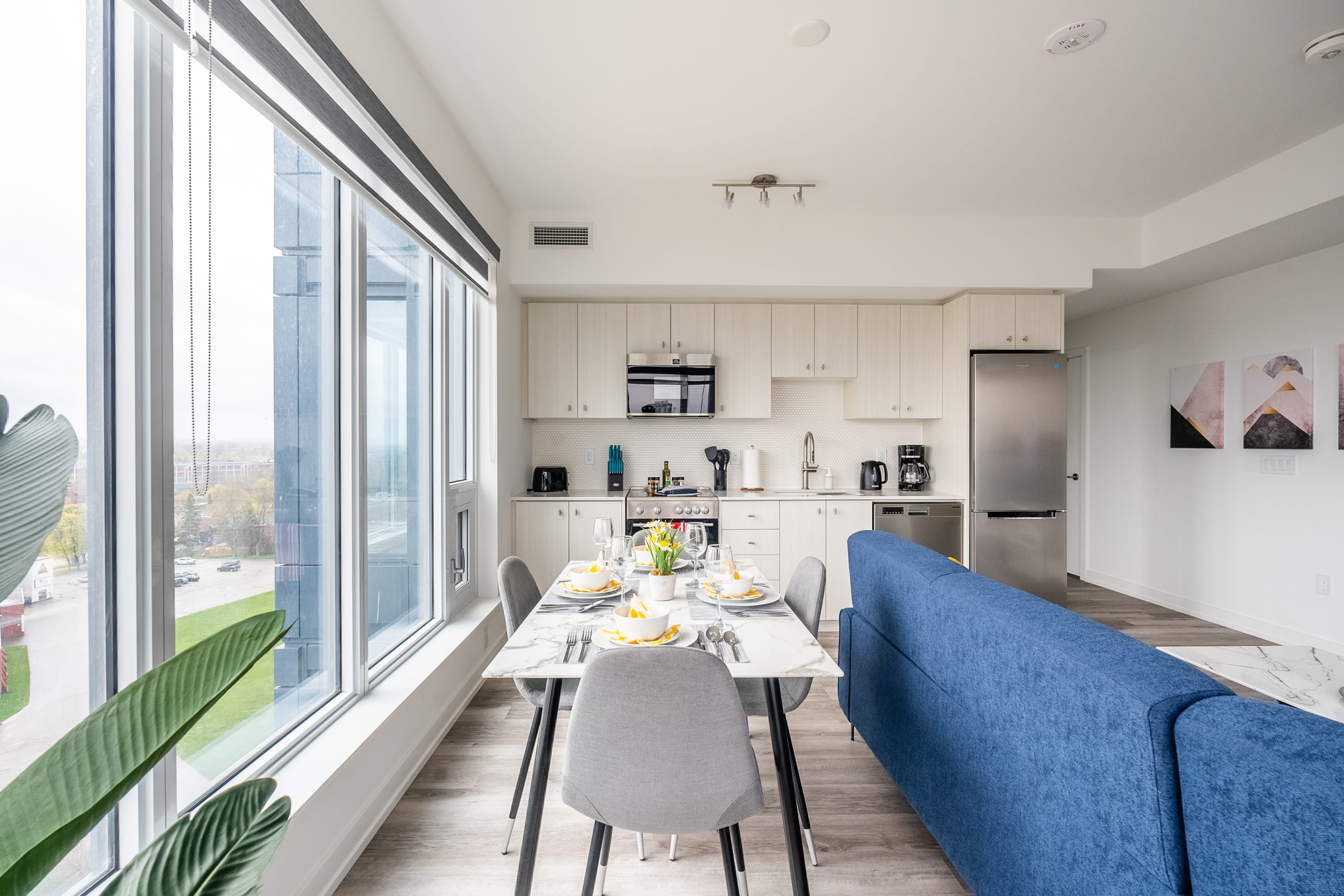 Modern kitchen with light wood cabinetry, white countertops, and stainless steel appliances, seamlessly flowing into a dining area with a marble-top table and gray chairs, all illuminated by large windows.