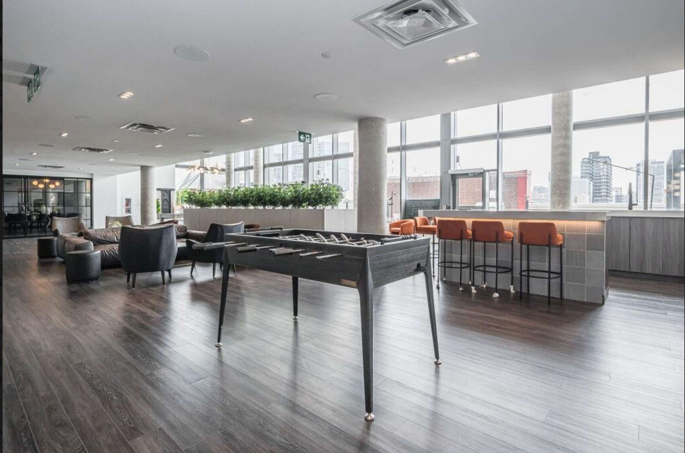 A modern and spacious common area with dark wood-look flooring, featuring a black foosball table in the foreground, a lounge area with grey sofas and armchairs, and a contemporary bar with orange bar stools and a light-colored countertop. Large windows offer city views in the background.
