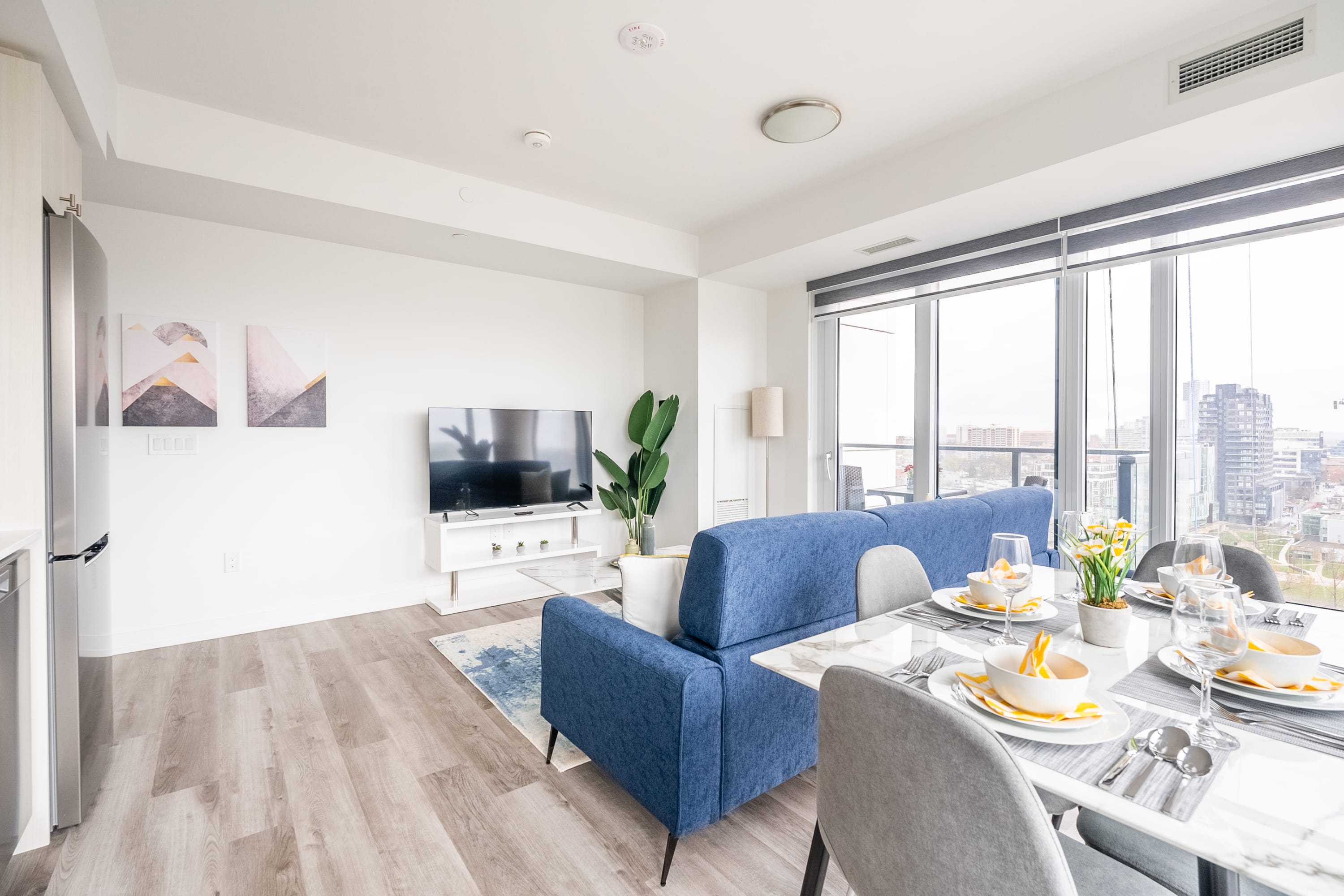 Contemporary living room with a large flat-screen TV on a white console, a blue sofa, and a dining area with a marble-top table and gray chairs. Large windows offer city views.