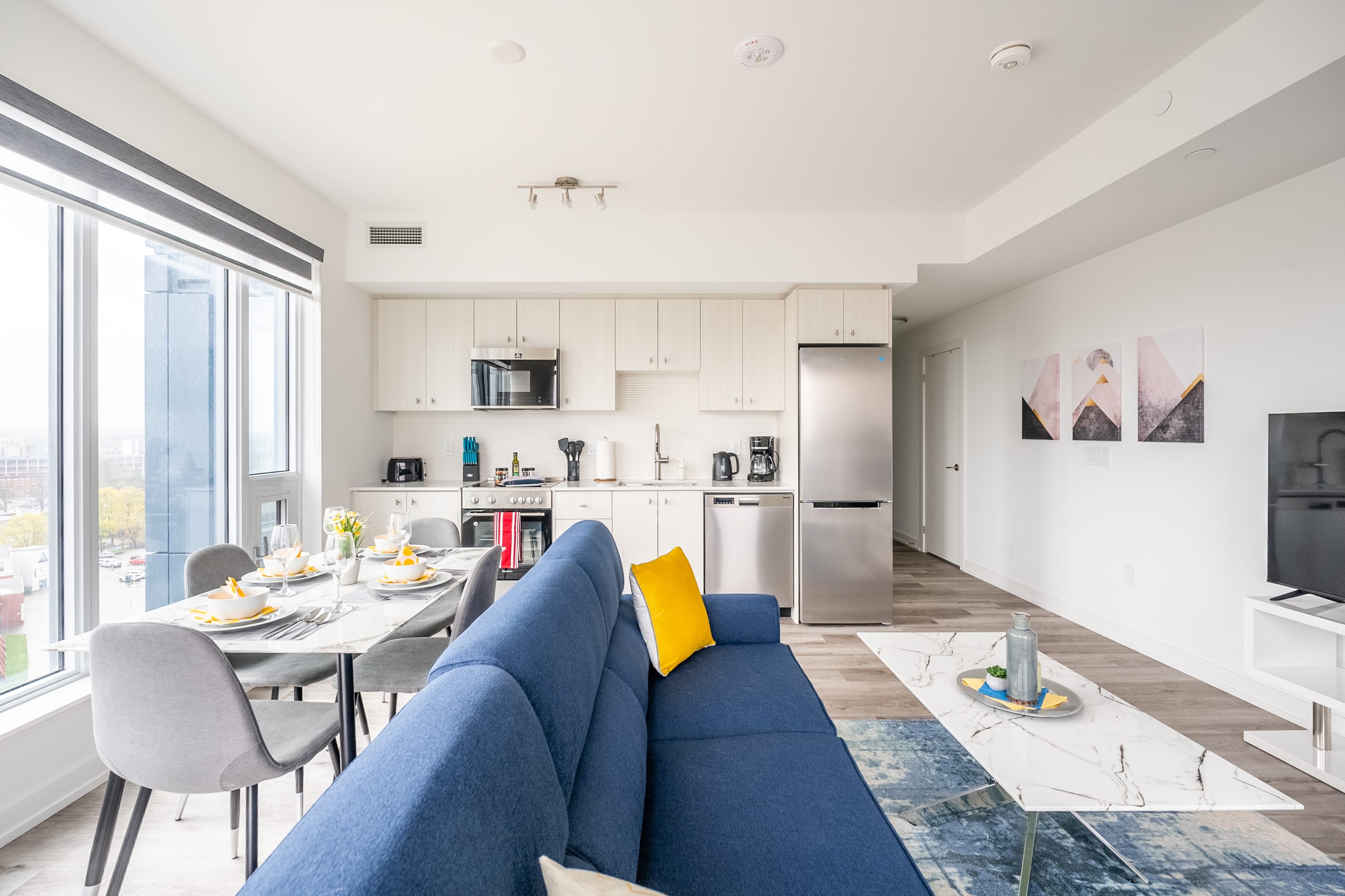 Bright open-concept living space featuring a blue sofa with a yellow accent pillow, a white marble coffee table, and a dining area with a set table, all illuminated by large windows overlooking a city view. The modern kitchen with light wood cabinets and stainless steel appliances is visible in the background.