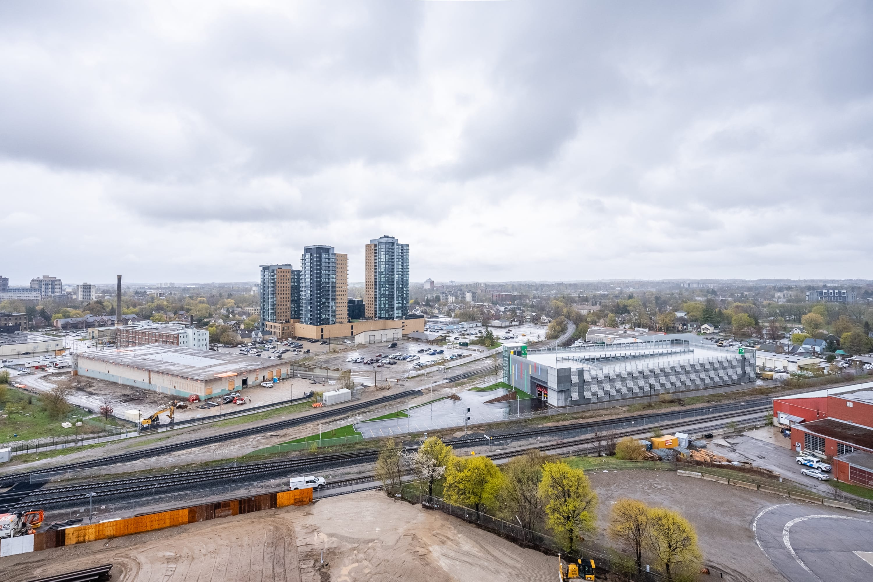 Panoramic view of a city landscape under an overcast sky, showing multiple high-rise buildings, industrial areas, railway tracks, and scattered green trees in the foreground.