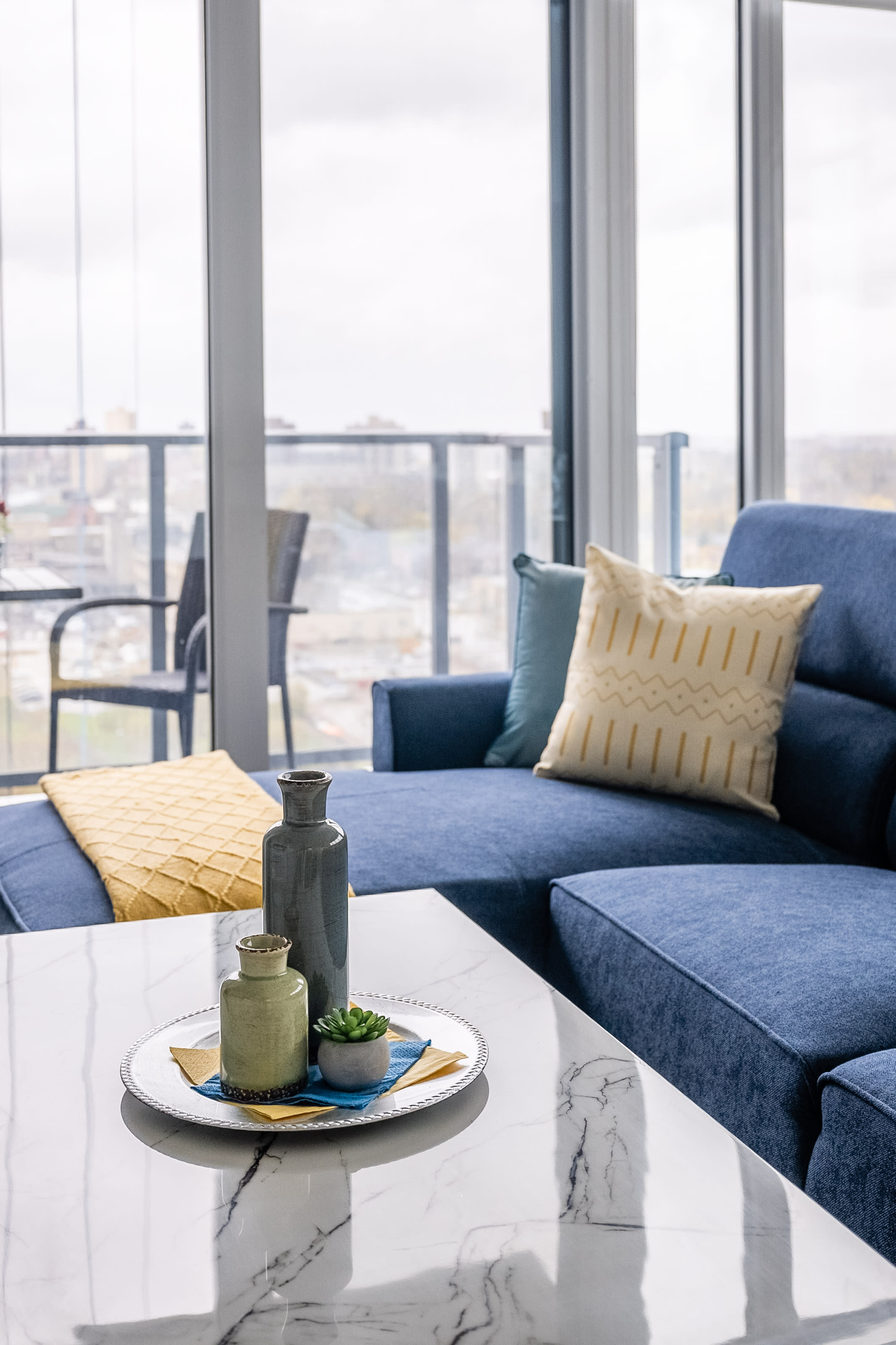 Close-up of a white marble coffee table adorned with decorative vases and a small succulent, positioned in front of a blue sectional sofa with yellow accent pillows. A balcony is visible through the large windows in the background.