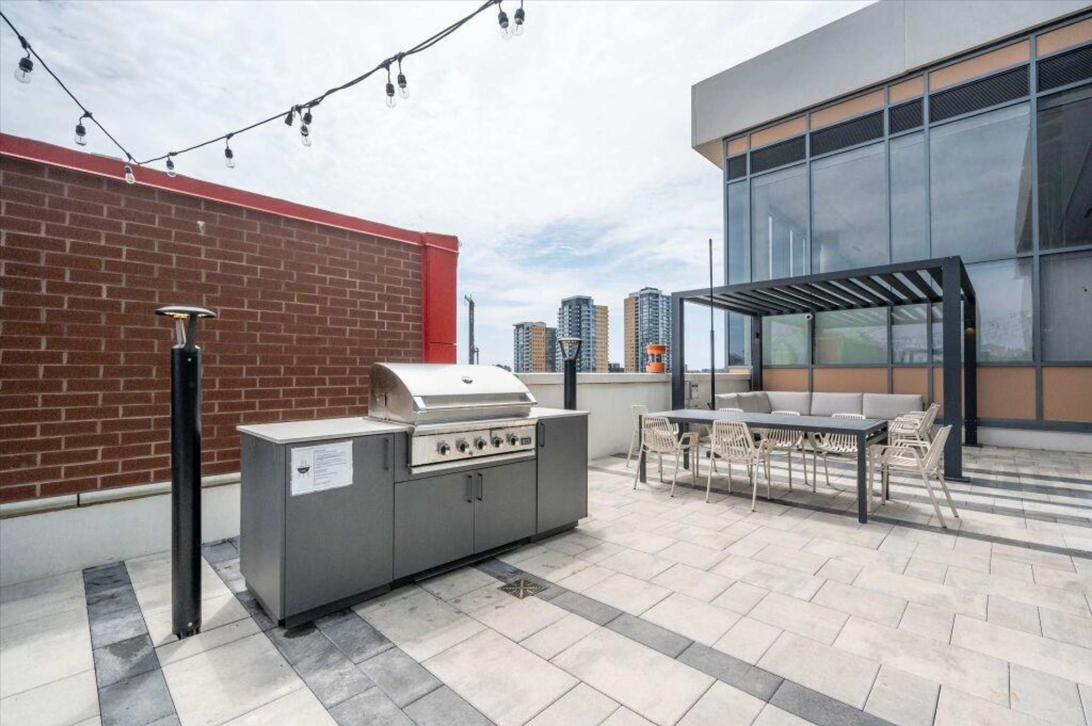 A spacious rooftop patio with light and dark grey patterned pavers, featuring a large stainless steel BBQ grill station, a long outdoor dining table with chairs under a modern black pergola, and string lights overhead. Tall buildings are visible in the distance under a cloudy sky.