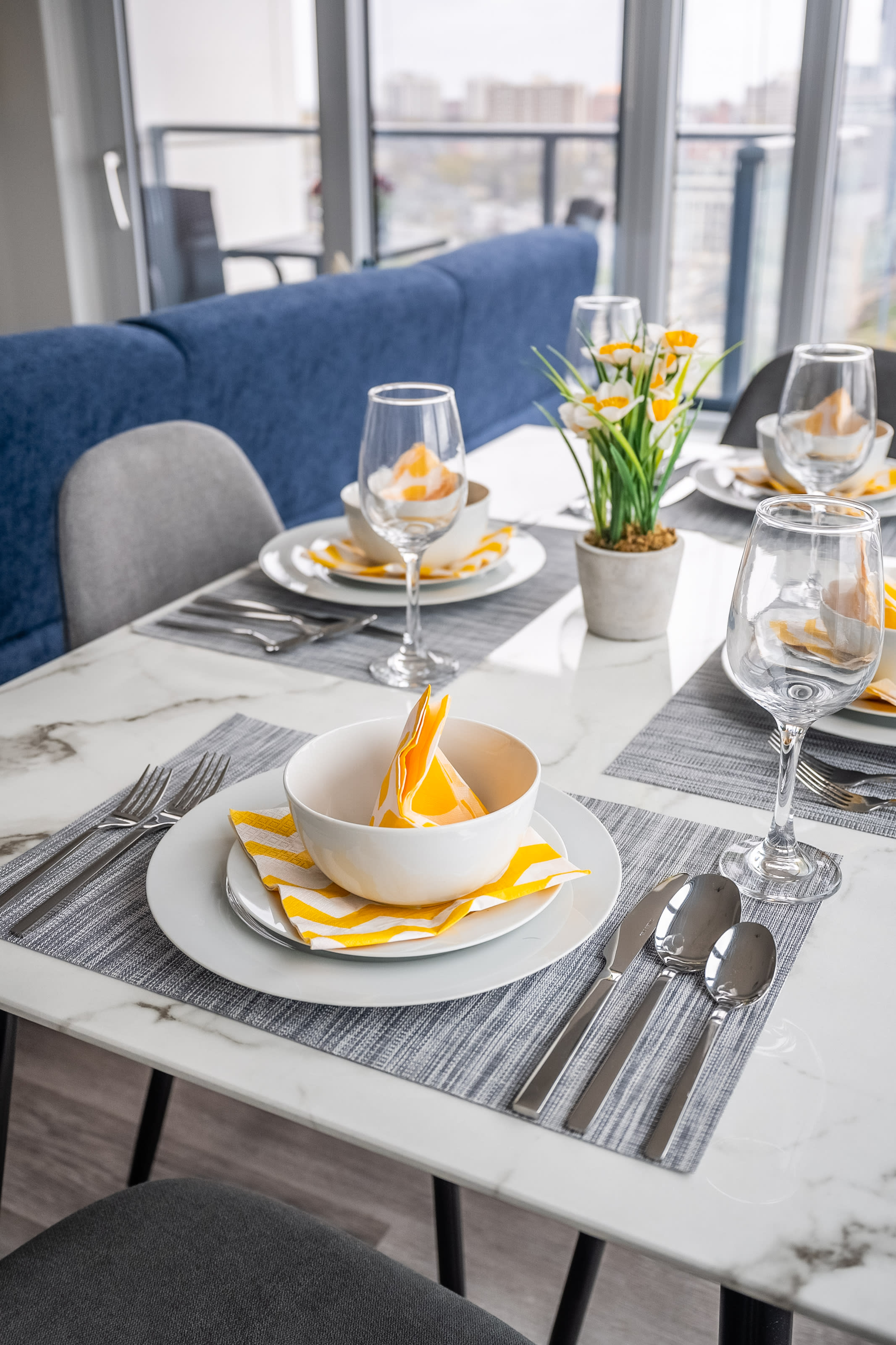 Close-up of a modern dining table set for four, featuring white plates, bowls with yellow napkins, wine glasses, and a small potted plant centerpiece, all on a white marble-patterned table with grey chairs.